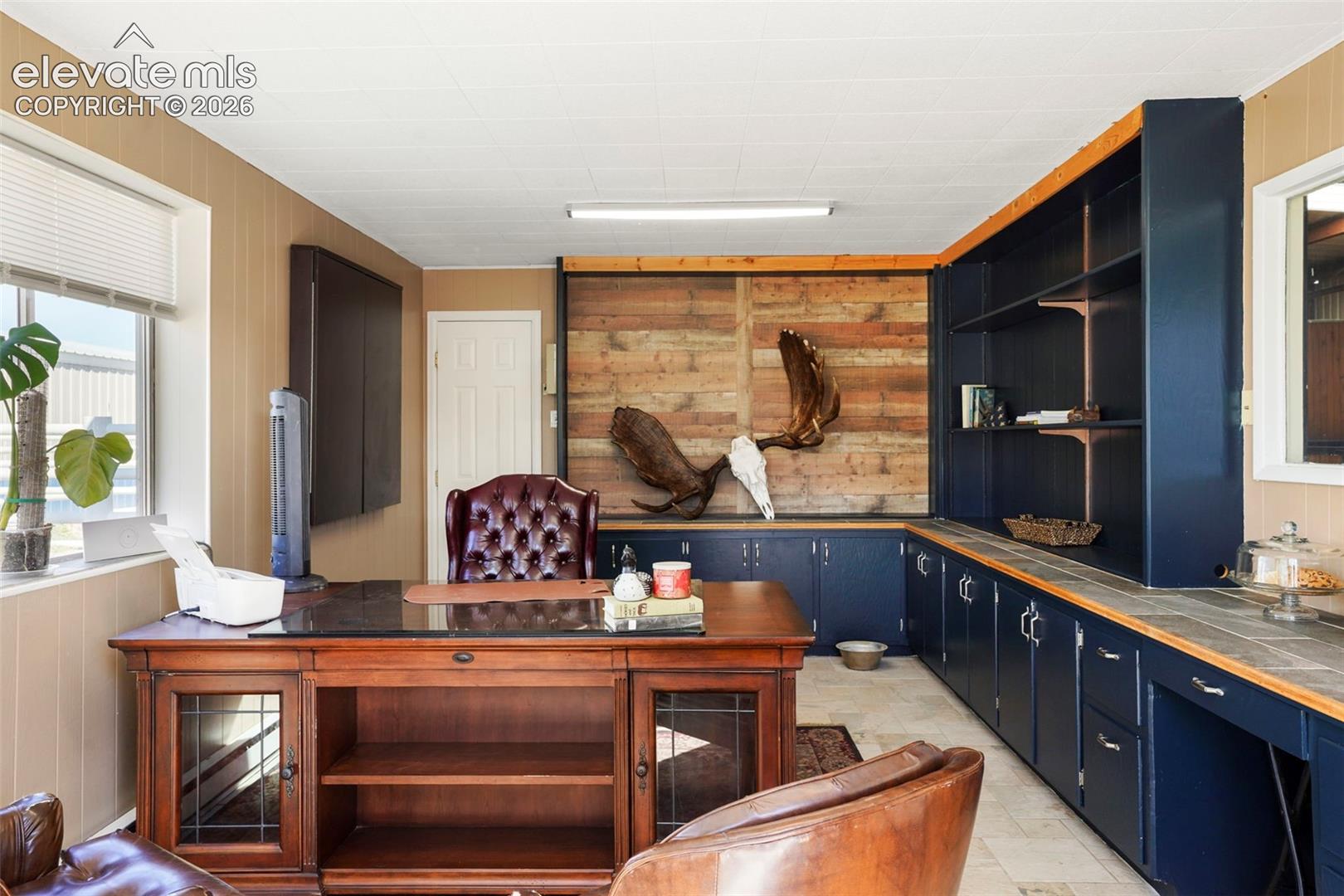 Custom built-in shelving, extensive built-in cabinetry with wood-finish countertops, wood-paneled accent wall, window with blinds, and durable tile flooring