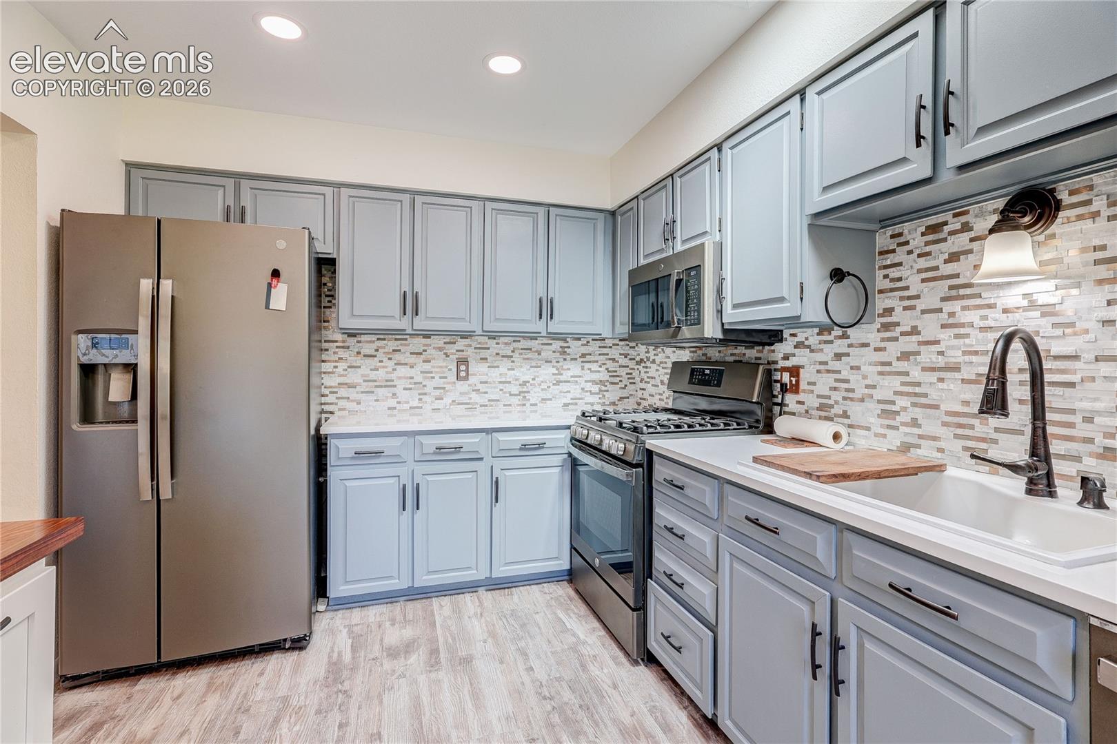 Kitchen featuring grey painted cabinetry, mosaic tile backsplash, stainless steel appliances, and a white apron-front sink with a high-arch pull-down faucet