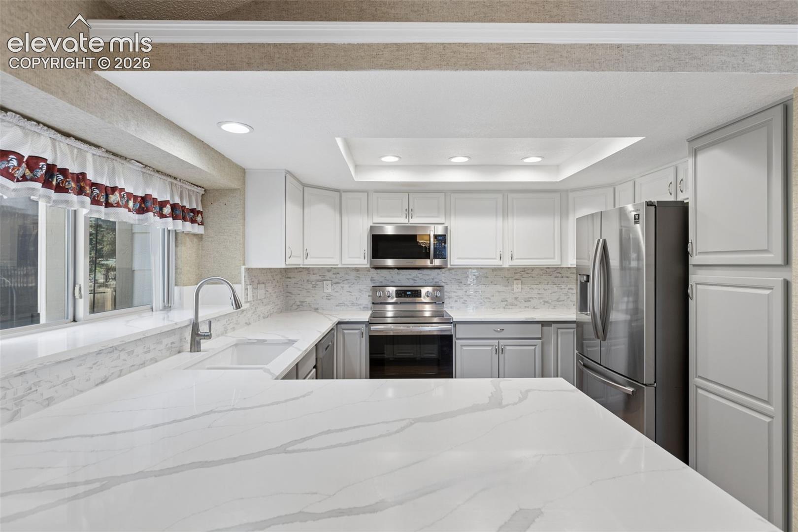 Kitchen featuring white cabinetry, stone-look countertops, stainless steel appliances, a tray ceiling with recessed lighting, and a tile backsplash
