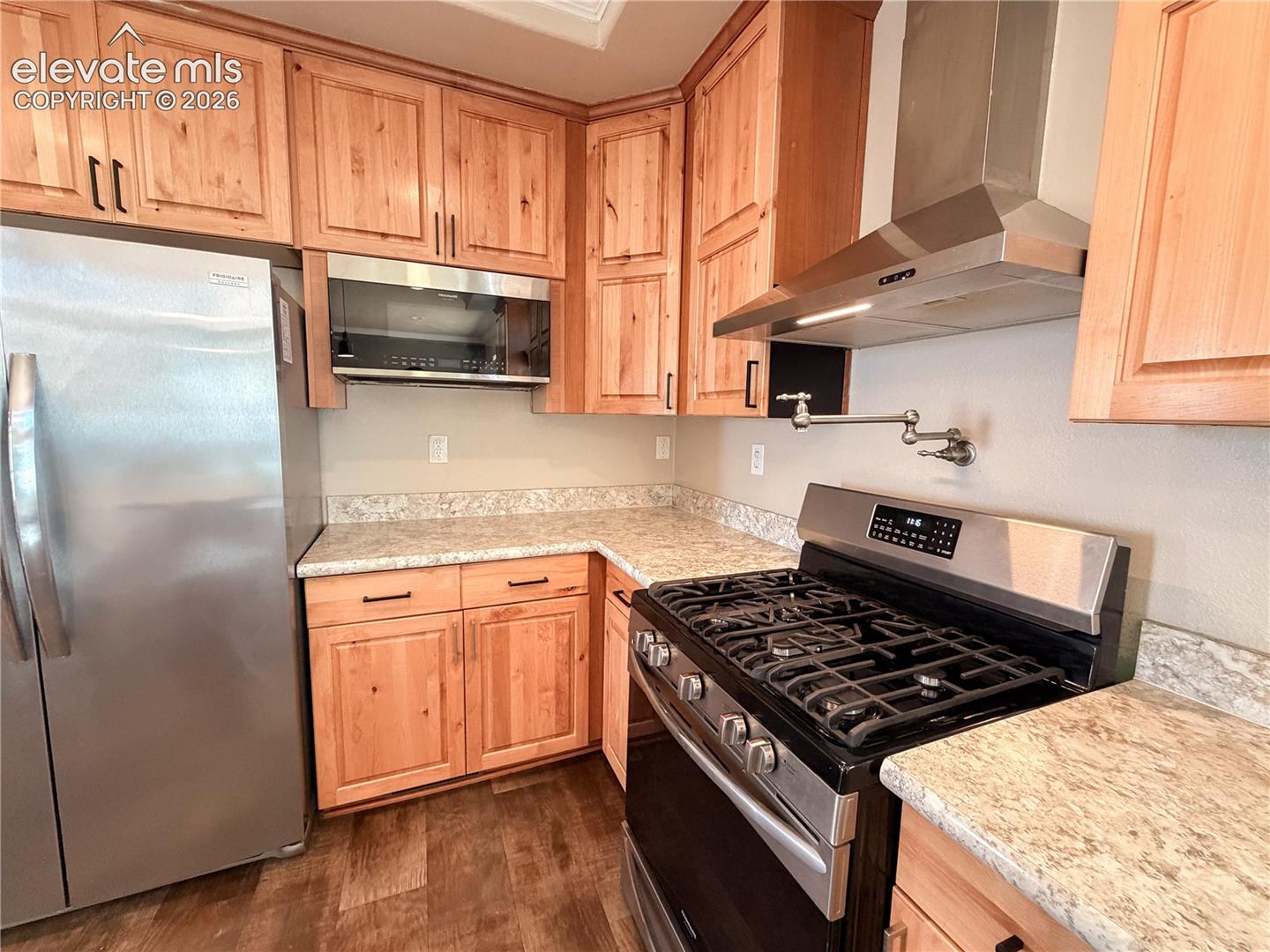 Kitchen featuring natural wood cabinetry, light-toned stone-finish countertops, stainless steel appliances, a wall-mounted pot filler, and a chimney-style range hood