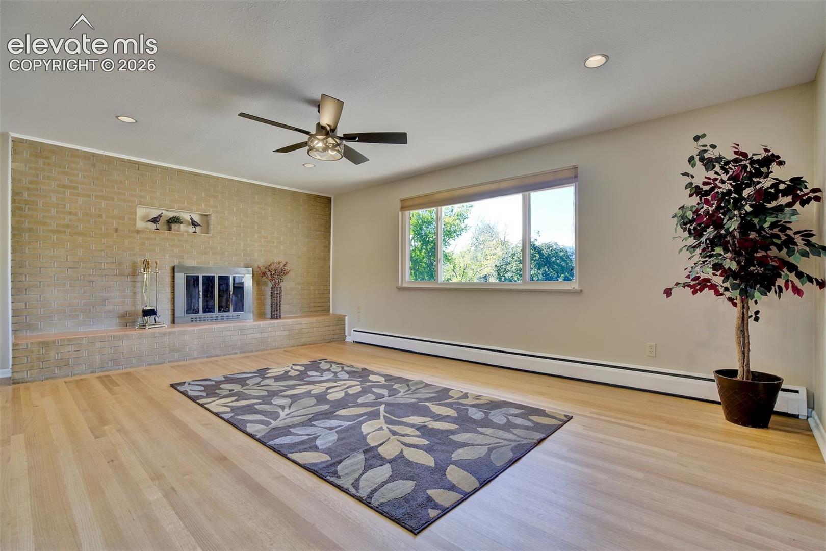 Living area featuring wood-finish flooring, a full-height brick fireplace with an integrated hearth, recessed ceiling lighting, and a multi-blade ceiling fan