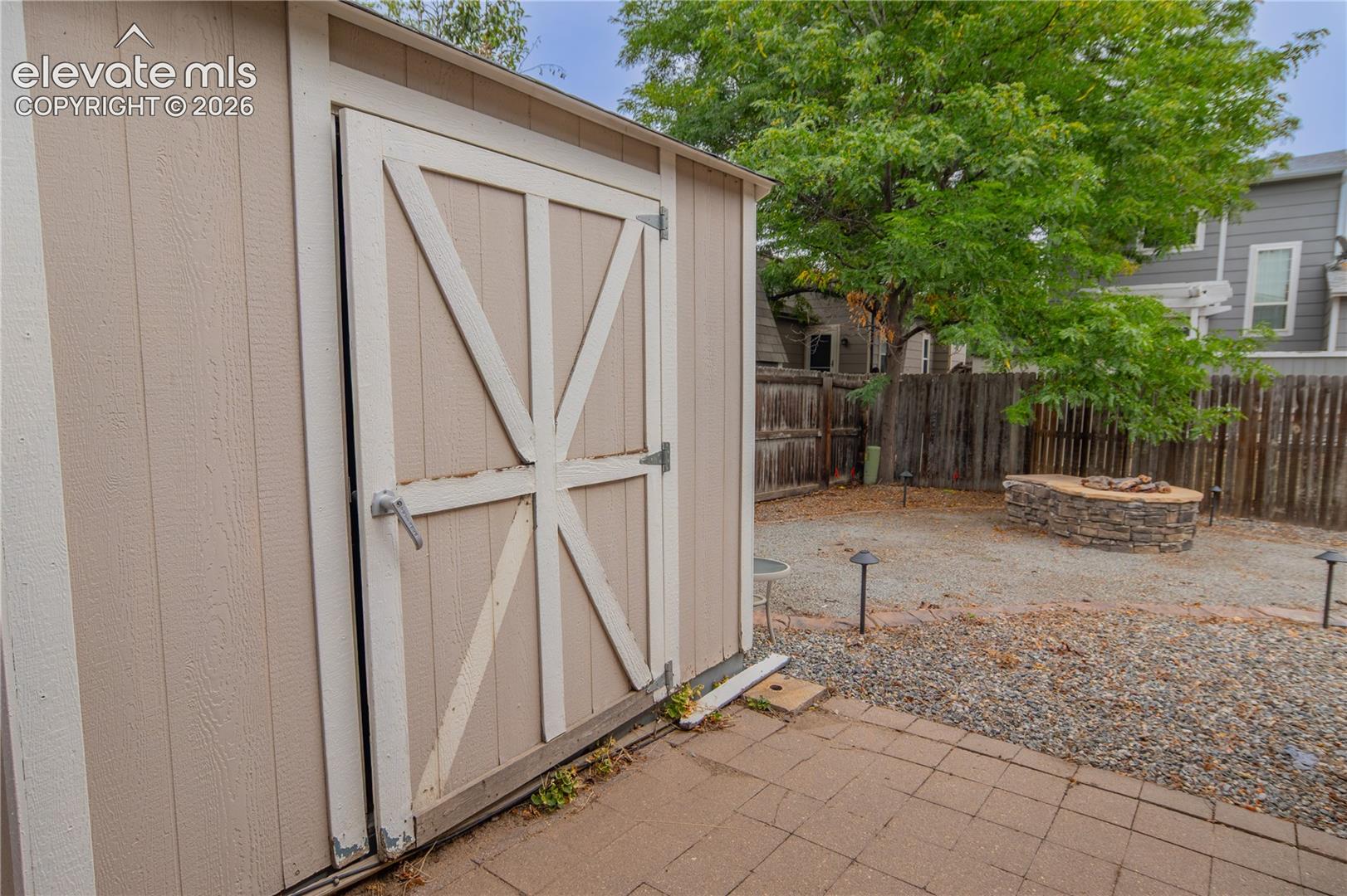 Outdoor storage shed with a panel door, paver patio, gravel yard, and a stone-built fire pit