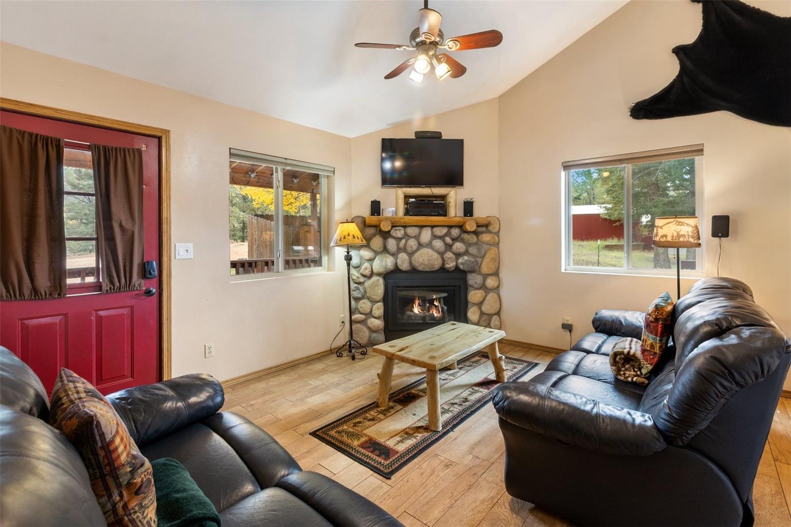 Vaulted ceiling with ceiling fan, stone-faced fireplace with wood mantel, wood-finish flooring, and a red entry door