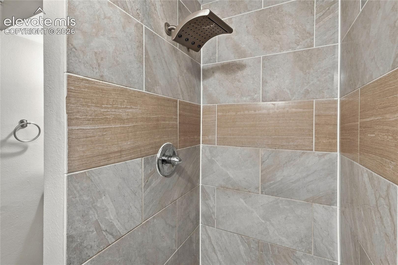 Basement shower featuring large format light gray tiles with a wood-finish accent band, a rain shower head, and a polished chrome control knob