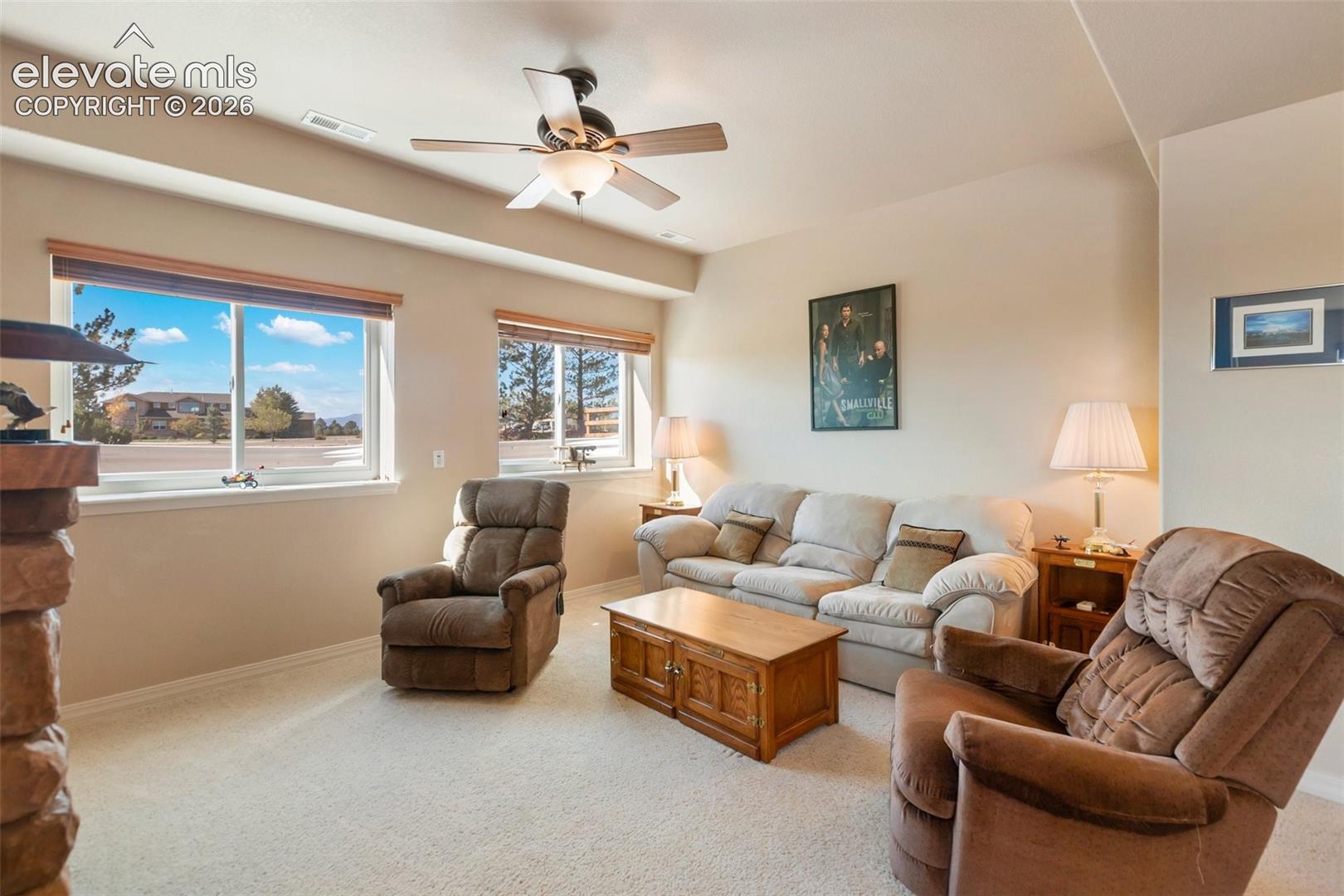 Living area featuring a textured stone fireplace, neutral wall-to-wall carpeting, dual windows with wood-tone blinds, and a ceiling-mounted light fixture with fan