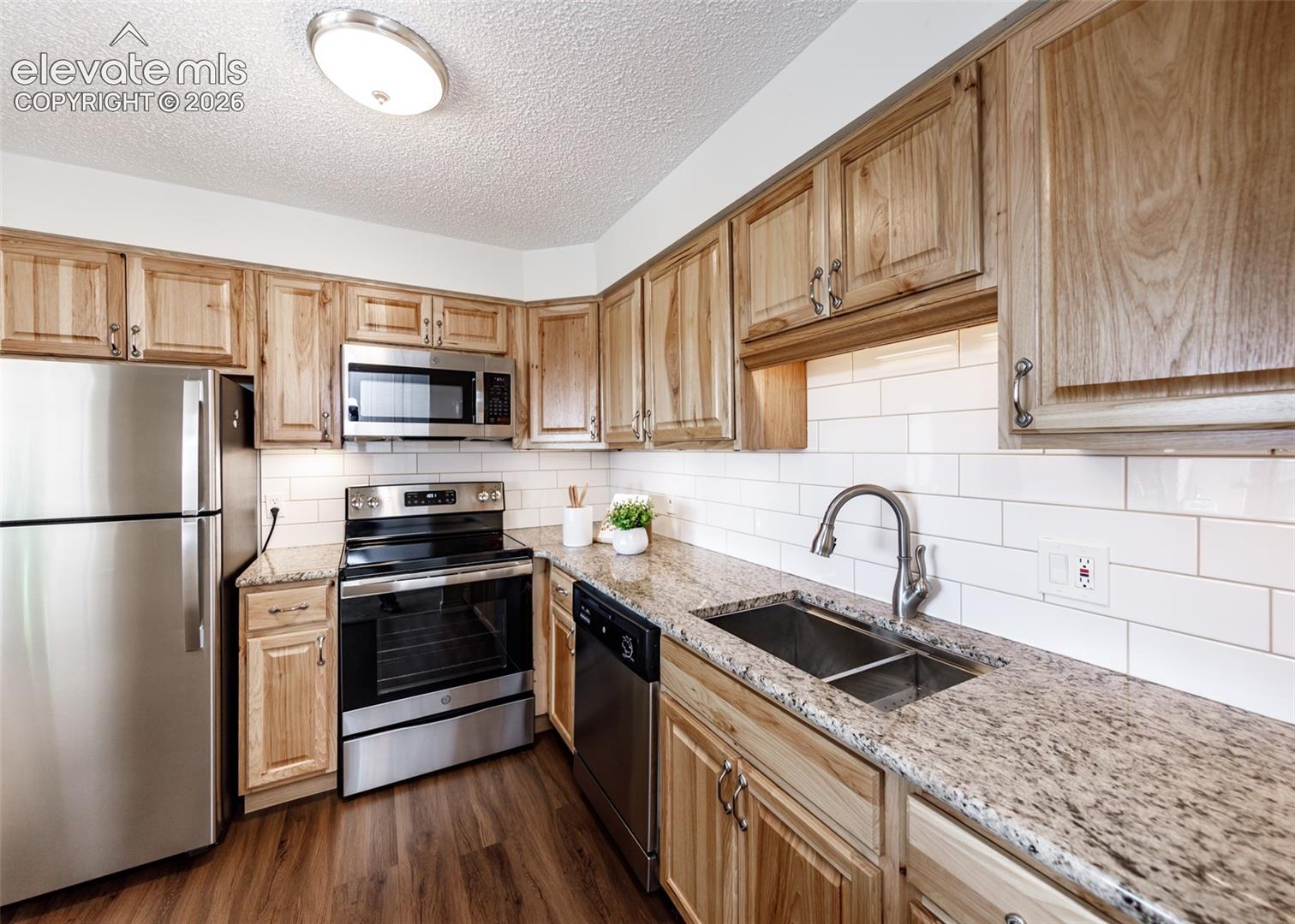 Kitchen featuring wood cabinetry, stone countertops, stainless steel appliances, subway tile backsplash, and wood-finish flooring