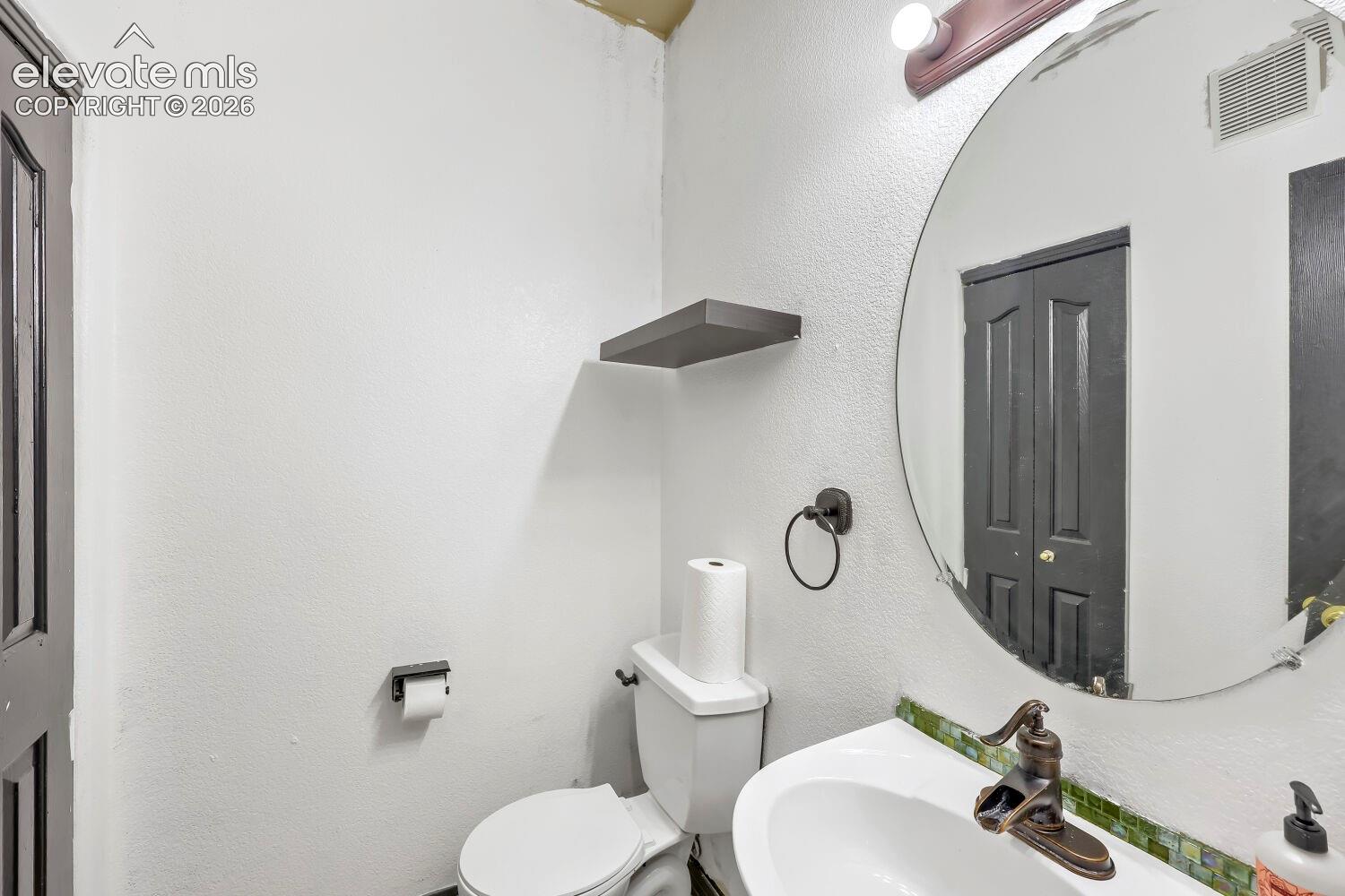 White ceramic toilet, pedestal sink with bronze-finish faucet, circular mirror, and wall-mounted shelving