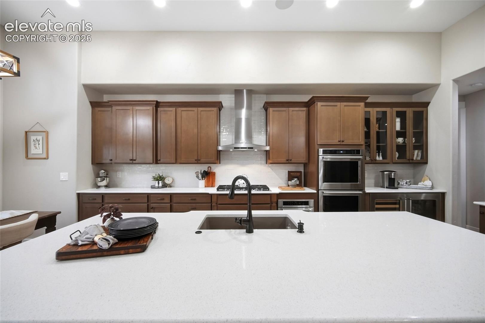 Kitchen featuring a quartz island countertop, dark wood-finish cabinetry, stainless steel double wall ovens, a stainless steel range hood, and a matte black gooseneck faucet
