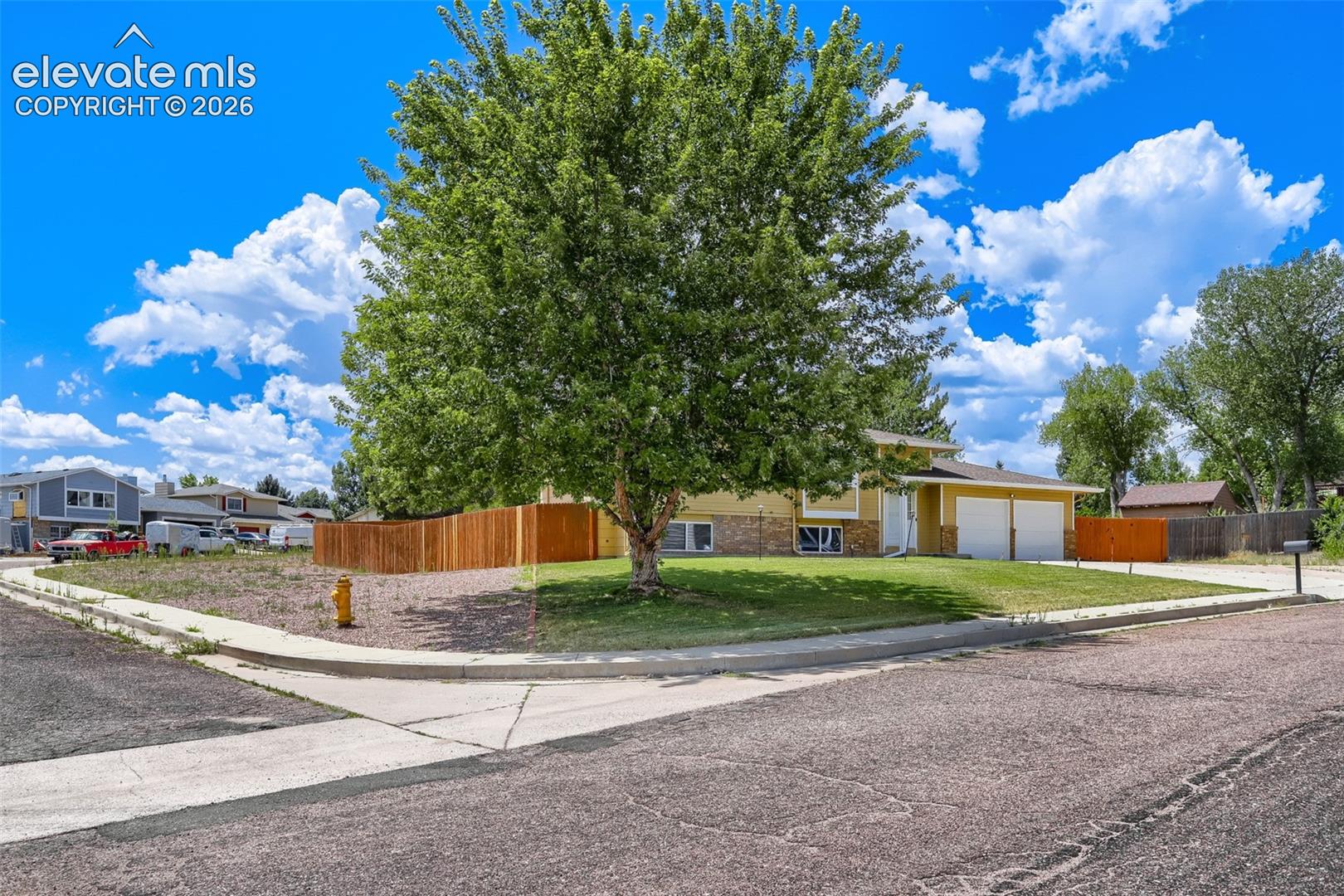 Split-level residence featuring a two-car garage, partial brick facade, and a mature front yard tree, corner lot.
