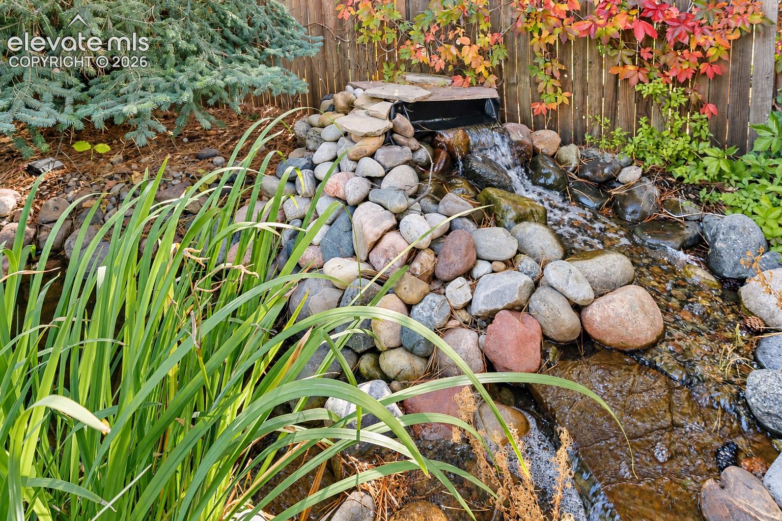 Custom rock water feature with tiered stones and integrated waterfall, surrounded by mature coniferous greenery and ornamental grasses