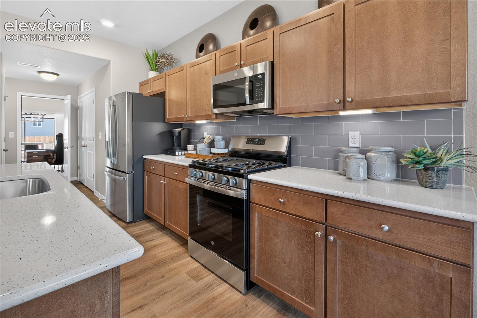 Kitchen featuring medium-toned wood cabinetry, stone-composite countertops, subway tile backsplash, stainless steel appliances, and wood-finish flooring