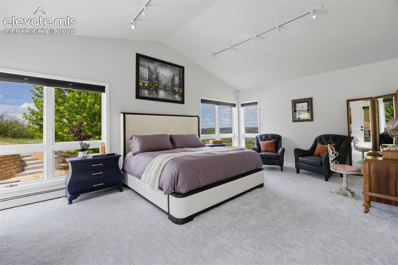 Primary suite with vaulted ceiling, walls of windows, plush carpet, sitting area, and walk-out to the patio.