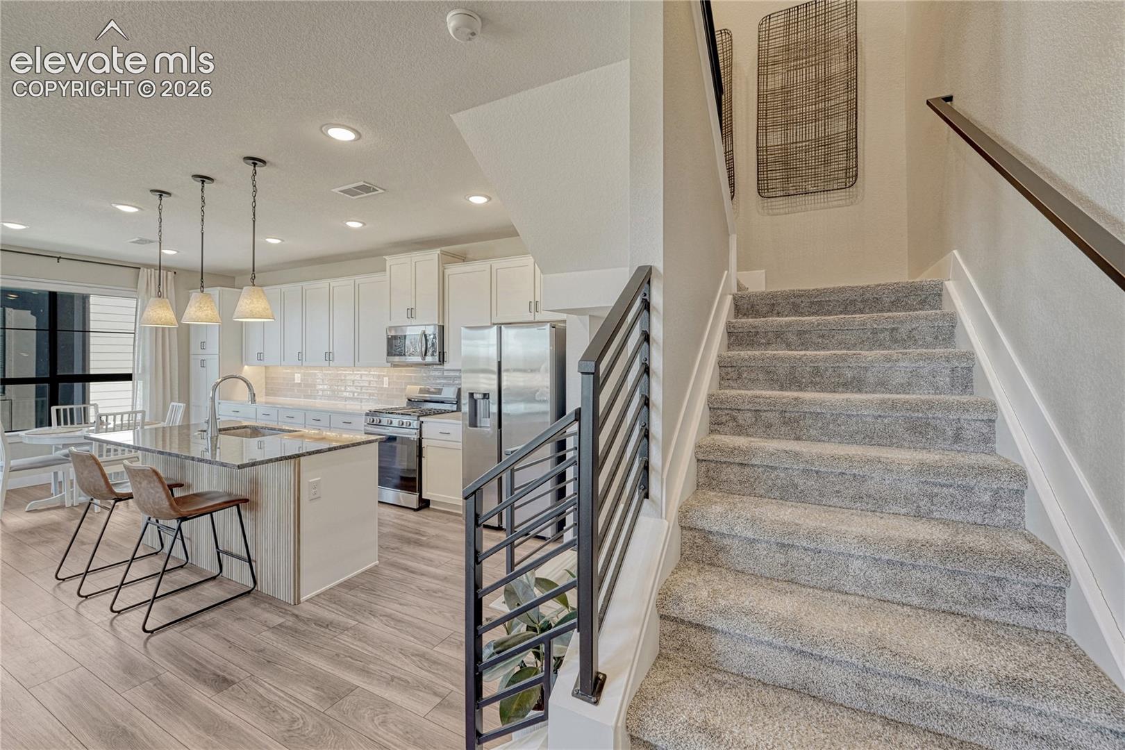 Open-concept interior featuring wood-finish flooring, a kitchen island with pendant lighting, white cabinetry, and a staircase with a black metal railing and carpeted steps