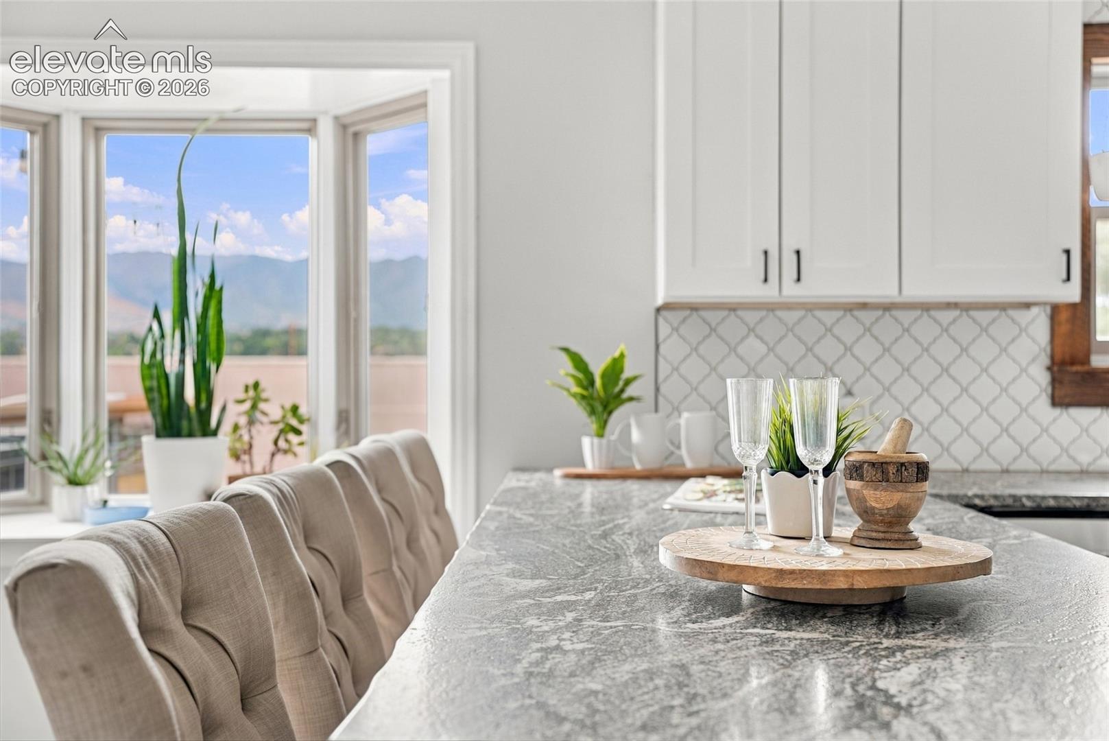 White shaker cabinetry with matte black hardware, gray patterned backsplash, bay window, natural wood-finish trim, and stone-finish countertops
