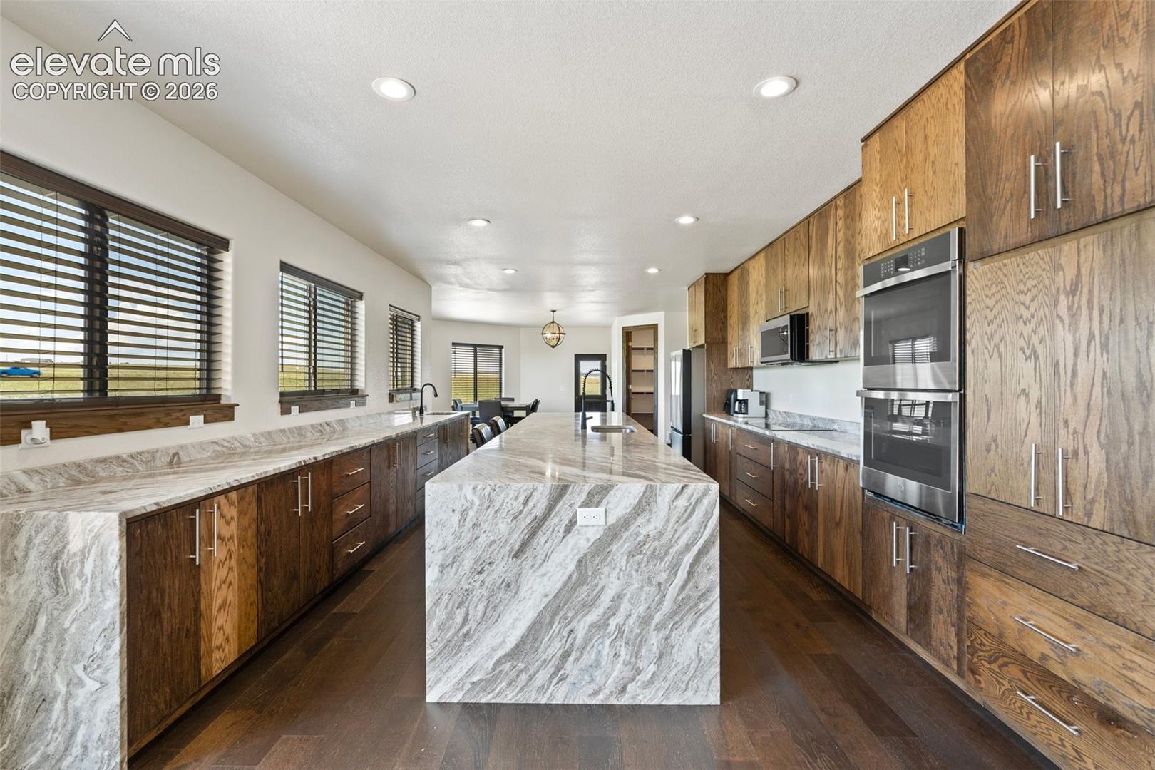 Spacious kitchen featuring a large waterfall island with integrated sink, extensive wood-finish cabinetry, stainless steel double ovens, and recessed lighting