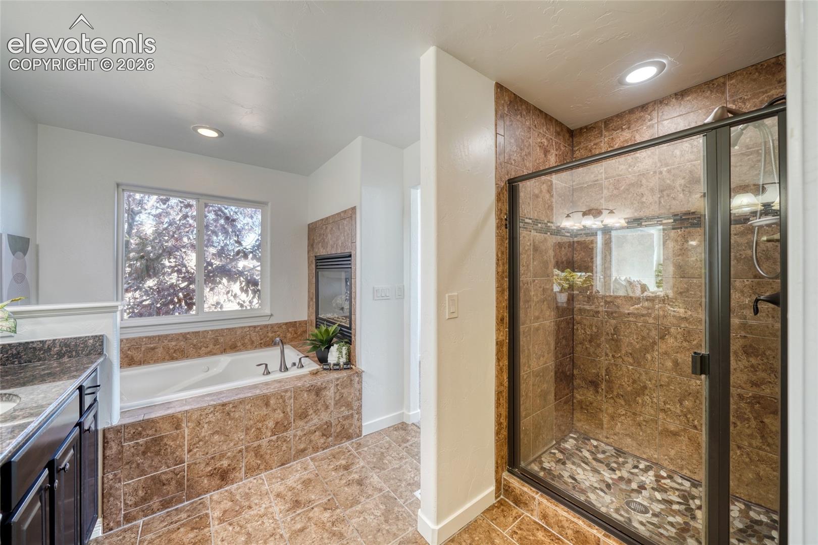 Built-in soaking tub with surrounding tile, separate shower enclosure with a pebble floor, and a double-pane window