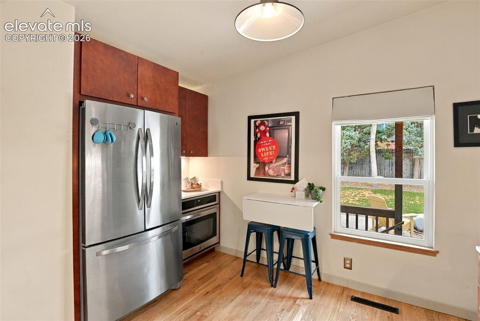 Kitchen featuring wood-finish cabinetry, stainless steel refrigerator, built-in oven, hardwood flooring, and a window with a fixed roller shade