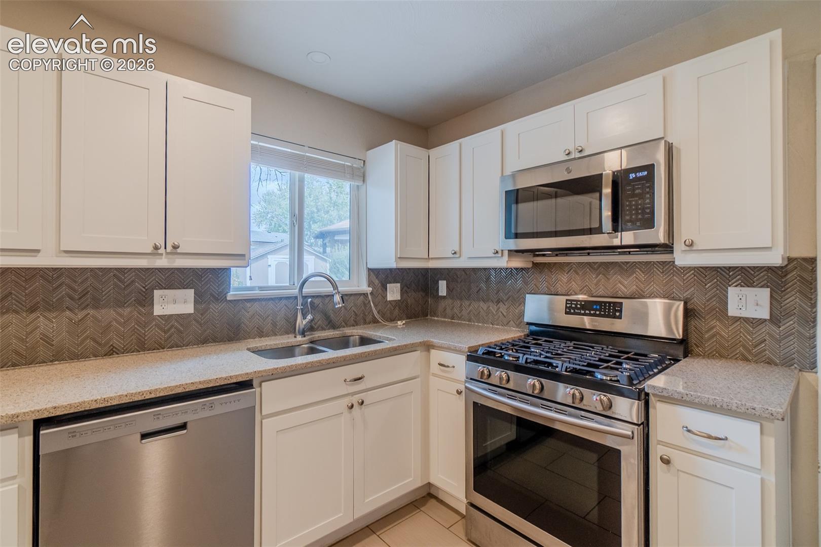 White shaker cabinetry, stainless steel appliances, patterned tile backsplash, dual basin undermount sink, and light-colored countertops