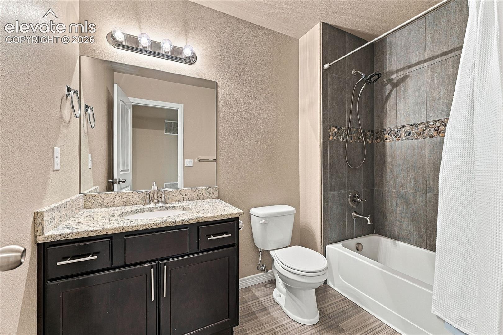 Dark-finish vanity cabinetry with a stone-composite countertop, wall-mounted four-bulb lighting fixture, and a tub-shower combination featuring slate-toned tile surround with a decorative stone mosaic accent