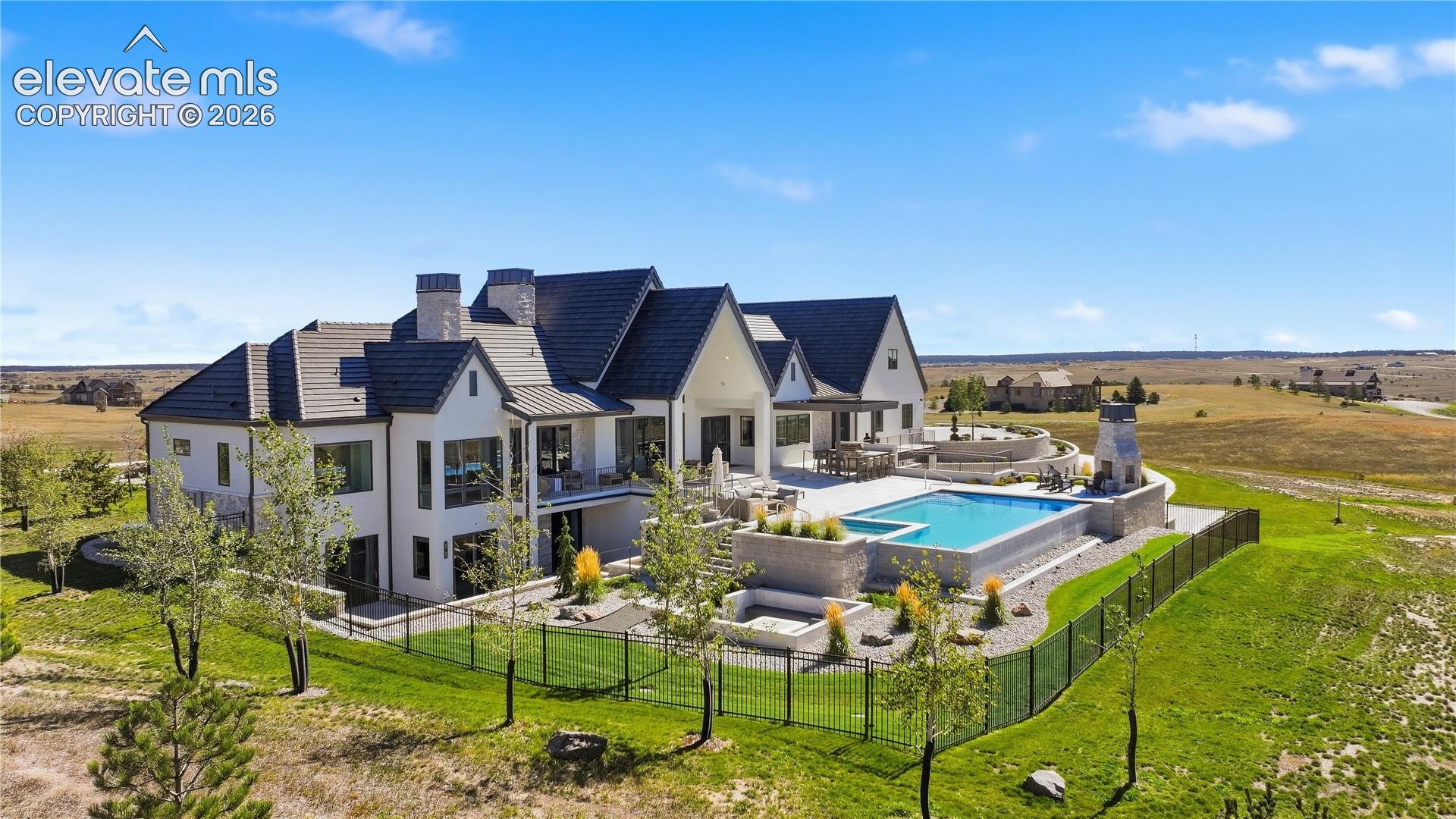 Multi-gabled estate featuring white stucco siding, a tiled roof, an elevated swimming pool with spa, a stone-clad outdoor fireplace, and tiered stone landscaping