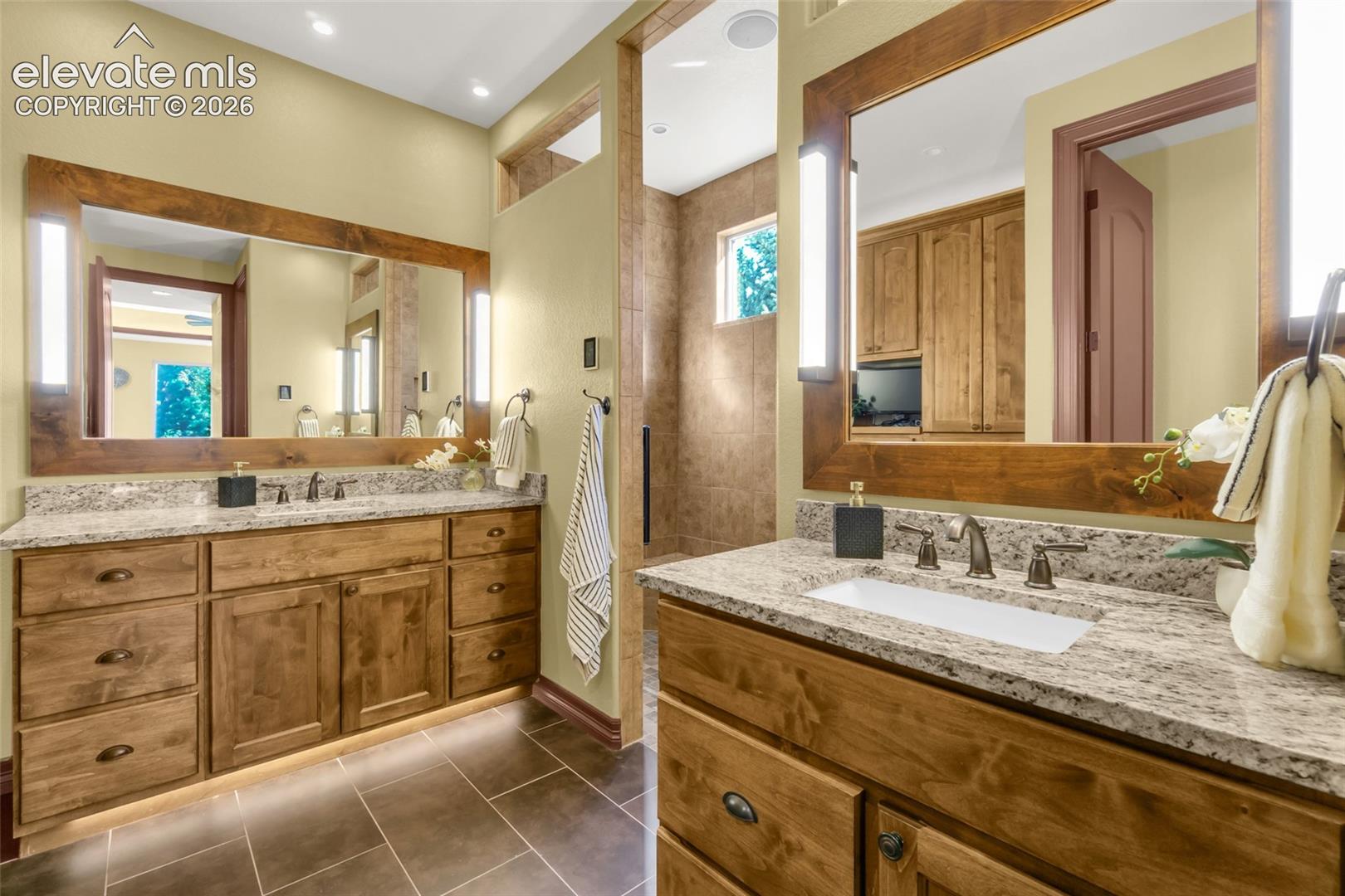 Dual-vanity bathroom featuring custom wood cabinetry, stone countertops with rectangular undermount sinks, wood-framed mirrors with integrated vertical lighting, and large-format tile flooring