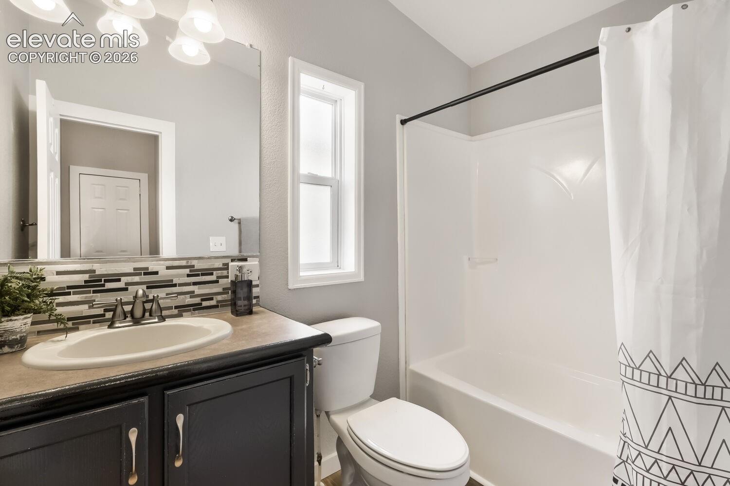 Bathroom featuring a vanity with dark cabinetry and a neutral-toned countertop, a multi-textured linear mosaic backsplash, a large frameless mirror, and a tub-shower combination with a surround