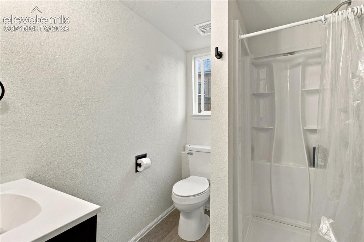 Bathroom featuring a single-basin vanity with a white countertop, wood-finish flooring, a porcelain toilet, and a molded shower surround with integrated shelving