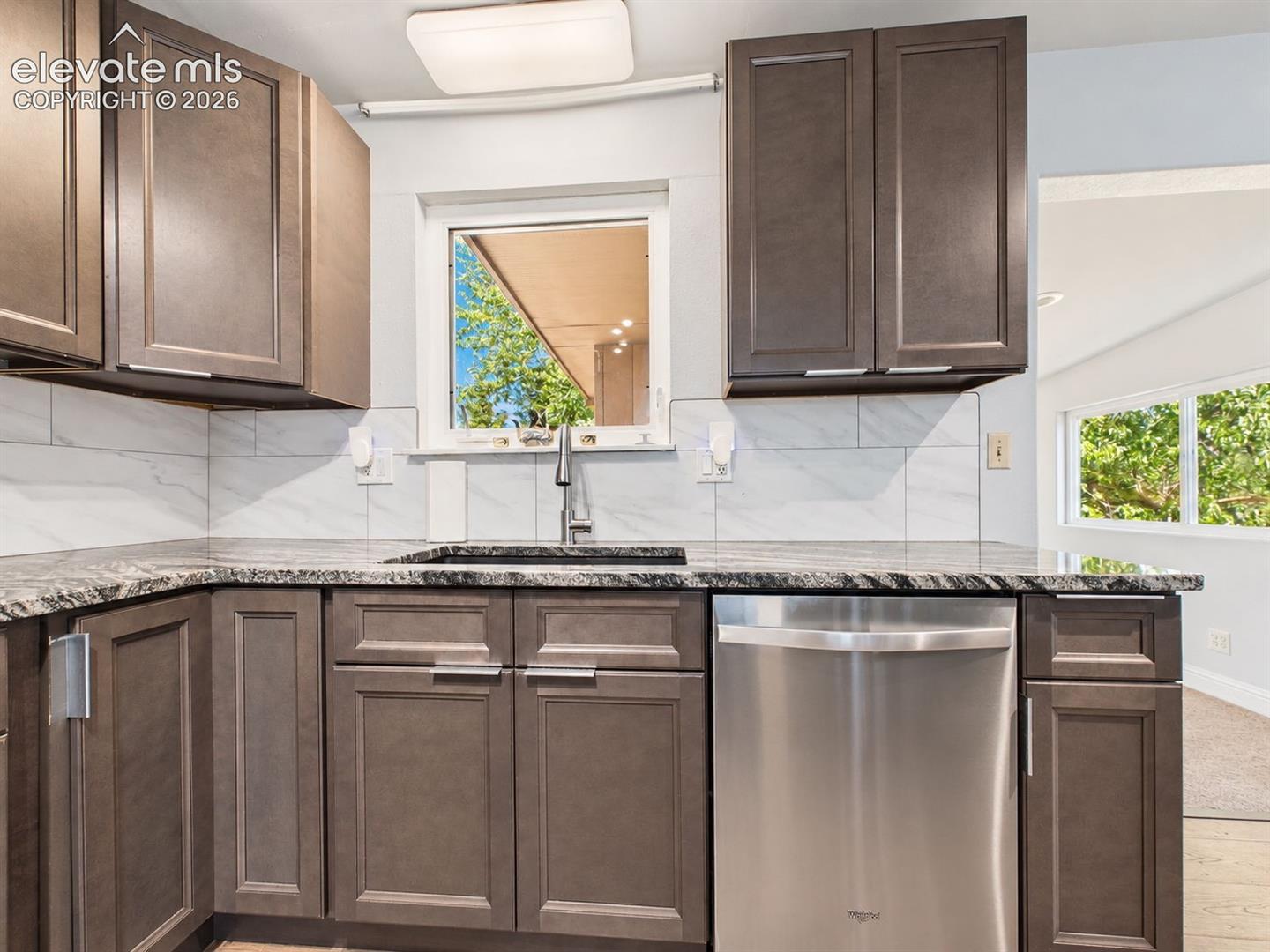 Sink-side view showcases abundant cabinetry, stainless steel dishwasher, tile backsplash, and a window over the sink bringing natural light into the workspace.