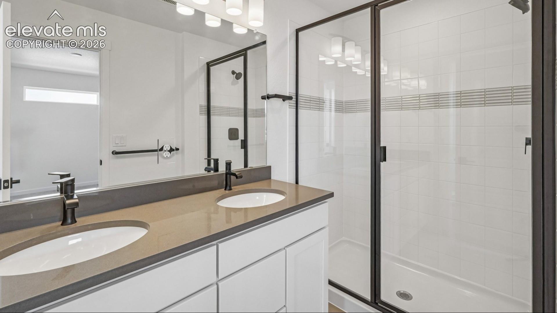 Bathroom featuring a dual-sink vanity with solid-surface countertops, dark metal fixtures, a glass-enclosed shower with tiled walls, and a full-width vanity mirror