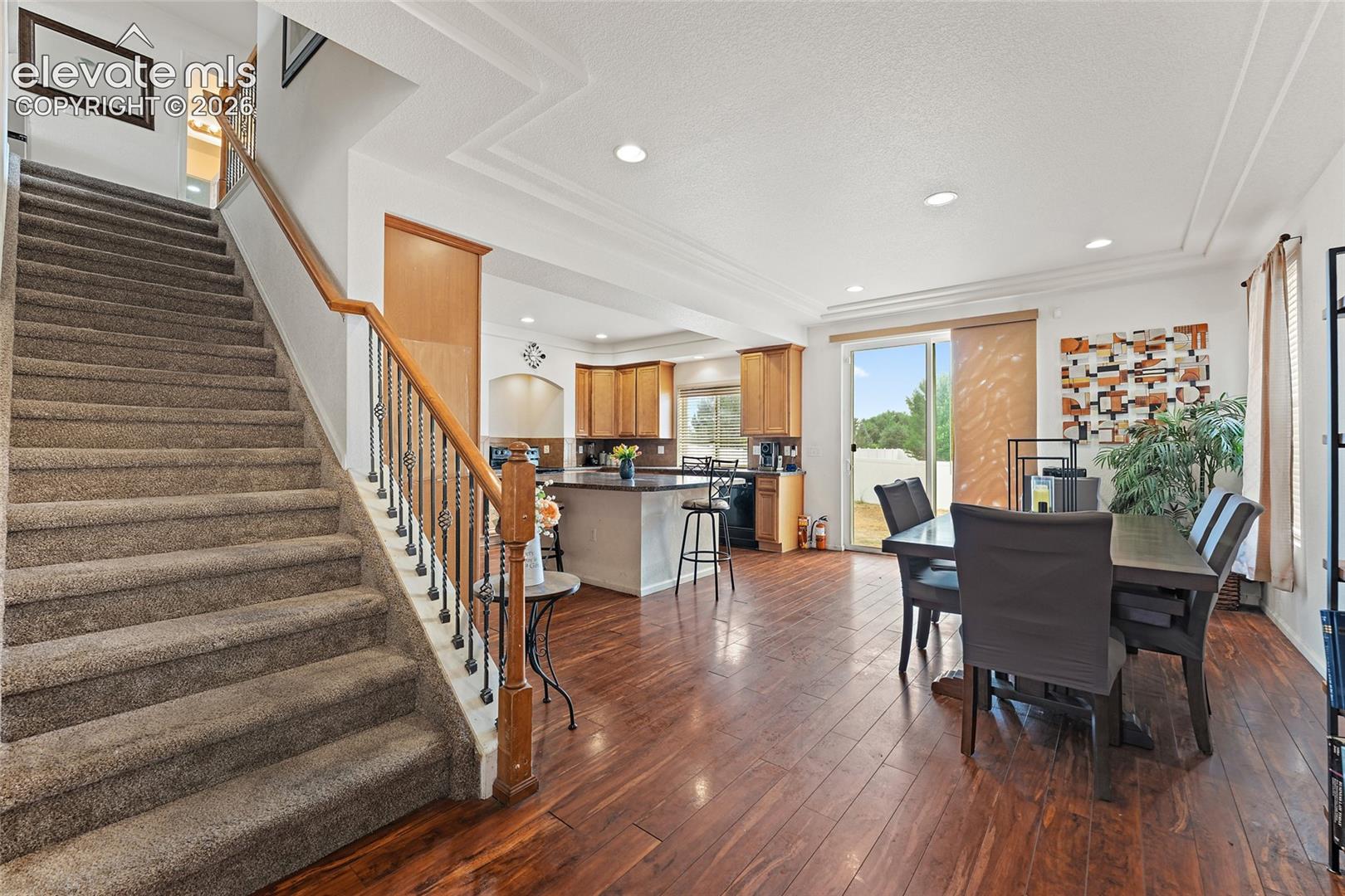 Staircase with carpeted steps and decorative iron balusters, kitchen featuring wood cabinetry and center island, and wood-finish flooring throughout the main level