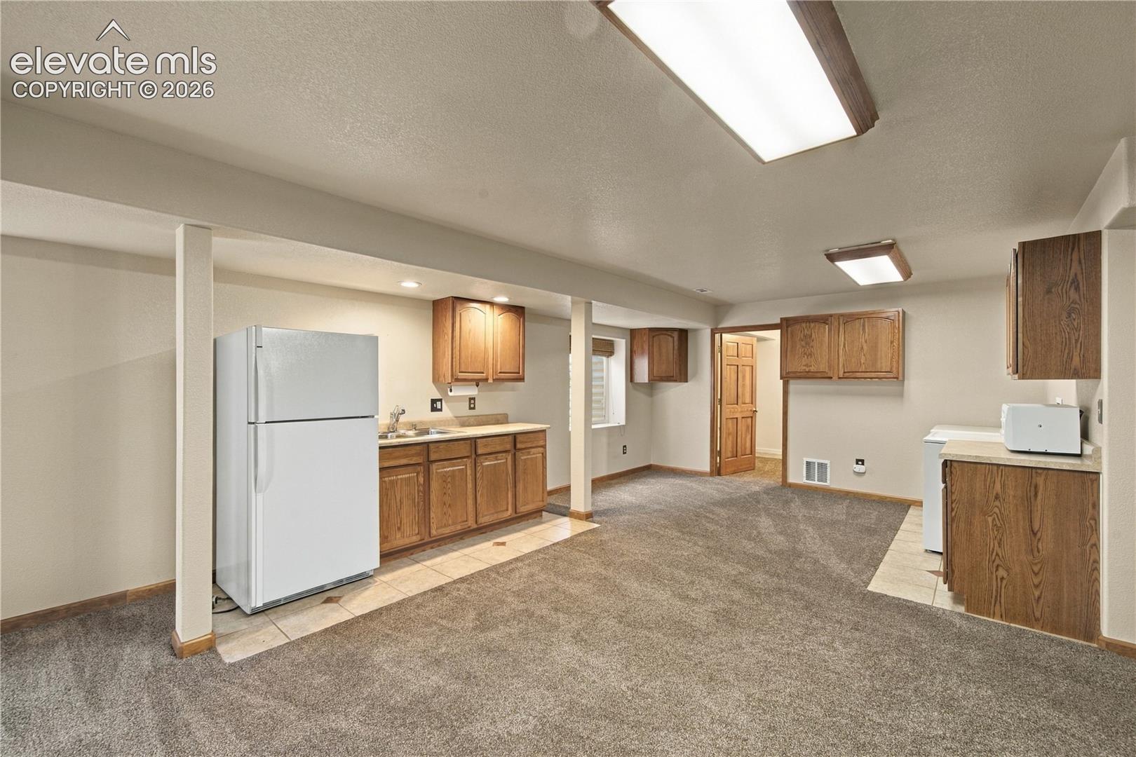 Kitchenette area featuring wood-finish cabinetry, neutral countertops, a stainless steel sink, tiled floor sections, and fluorescent recessed lighting