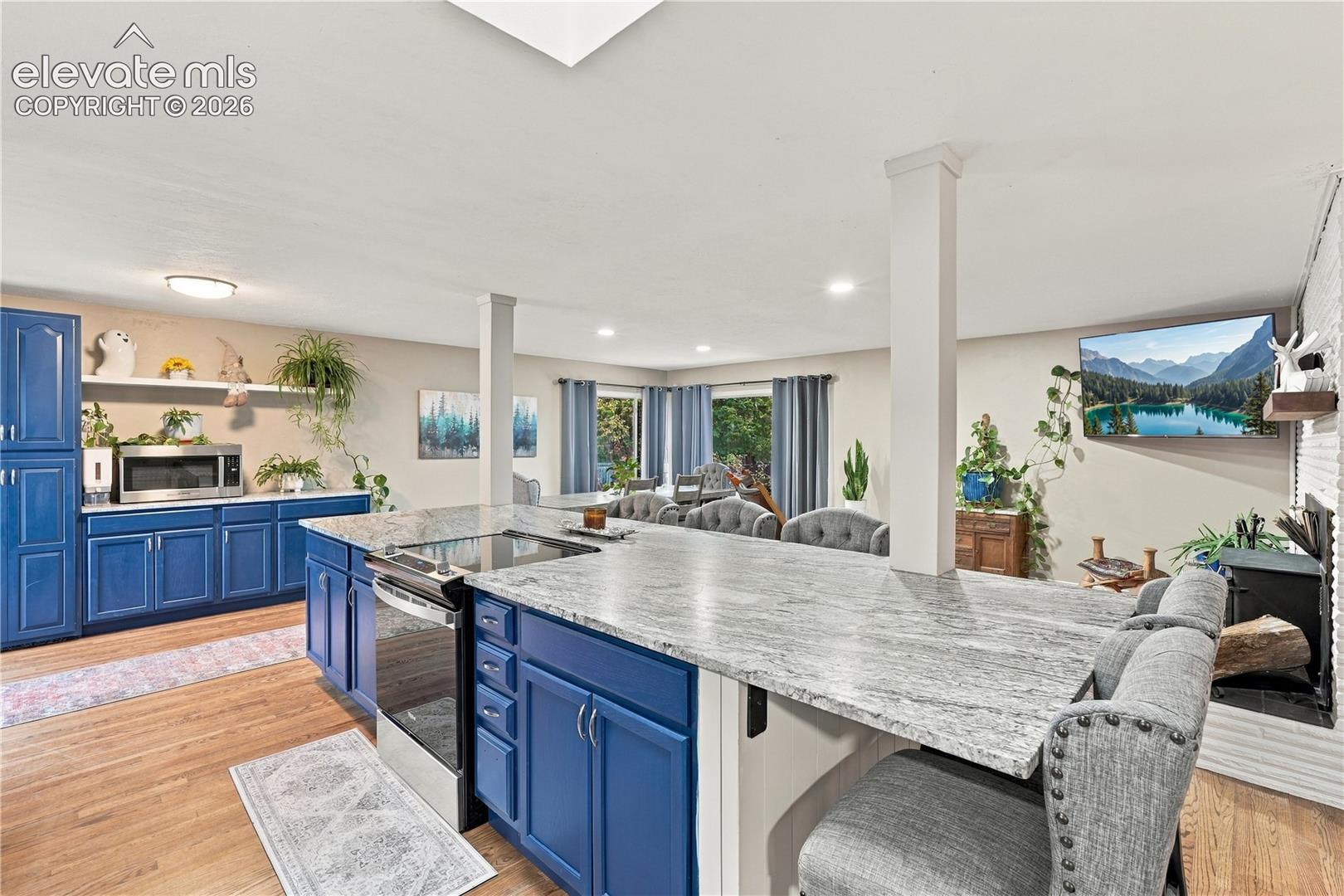 Spacious kitchen featuring blue cabinetry, stone-finish countertops, stainless steel slide-in range, wood-finish flooring, and a white brick fireplace surround