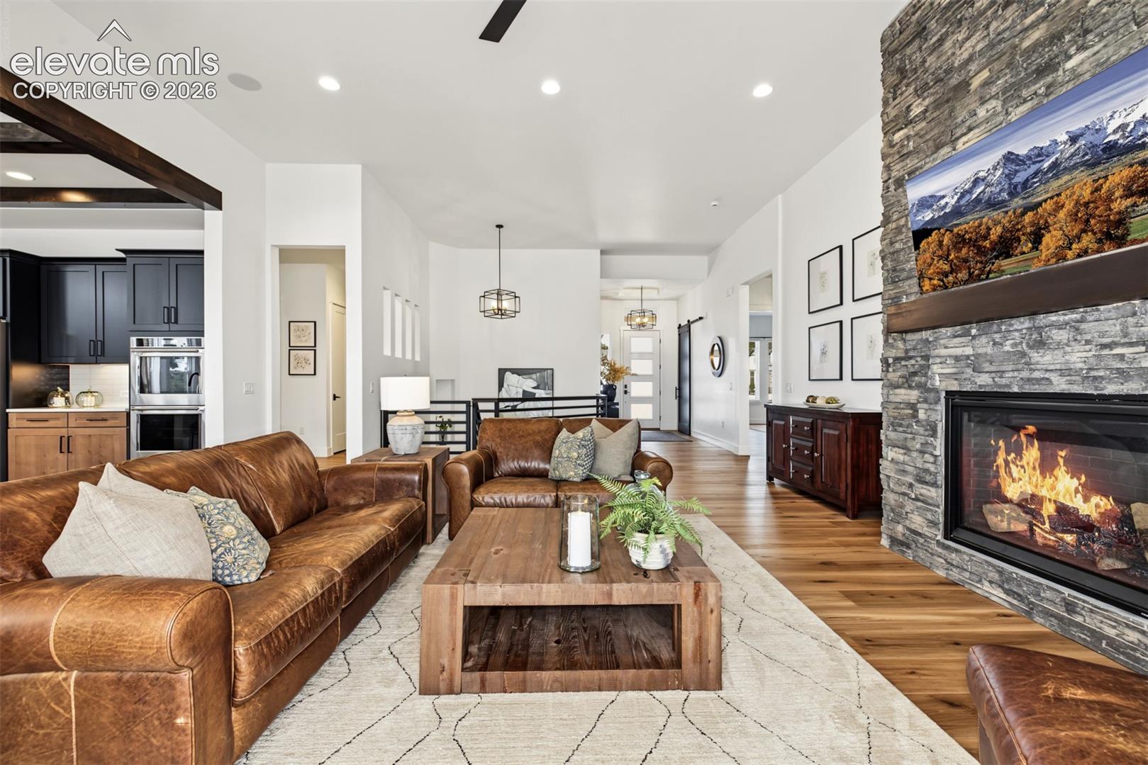 Stunning great room featuring soaring ceilings, expansive windows, and a dramatic floor-to-ceiling stone fireplace. Beautiful wood flooring and an open-concept design create a warm, inviting living space.