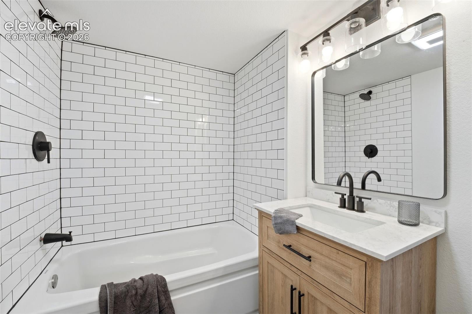 Bathroom featuring a white subway tile surround, matte black fixtures, a single vanity with wood-finish cabinetry, and a rectangular mirror with integrated lighting