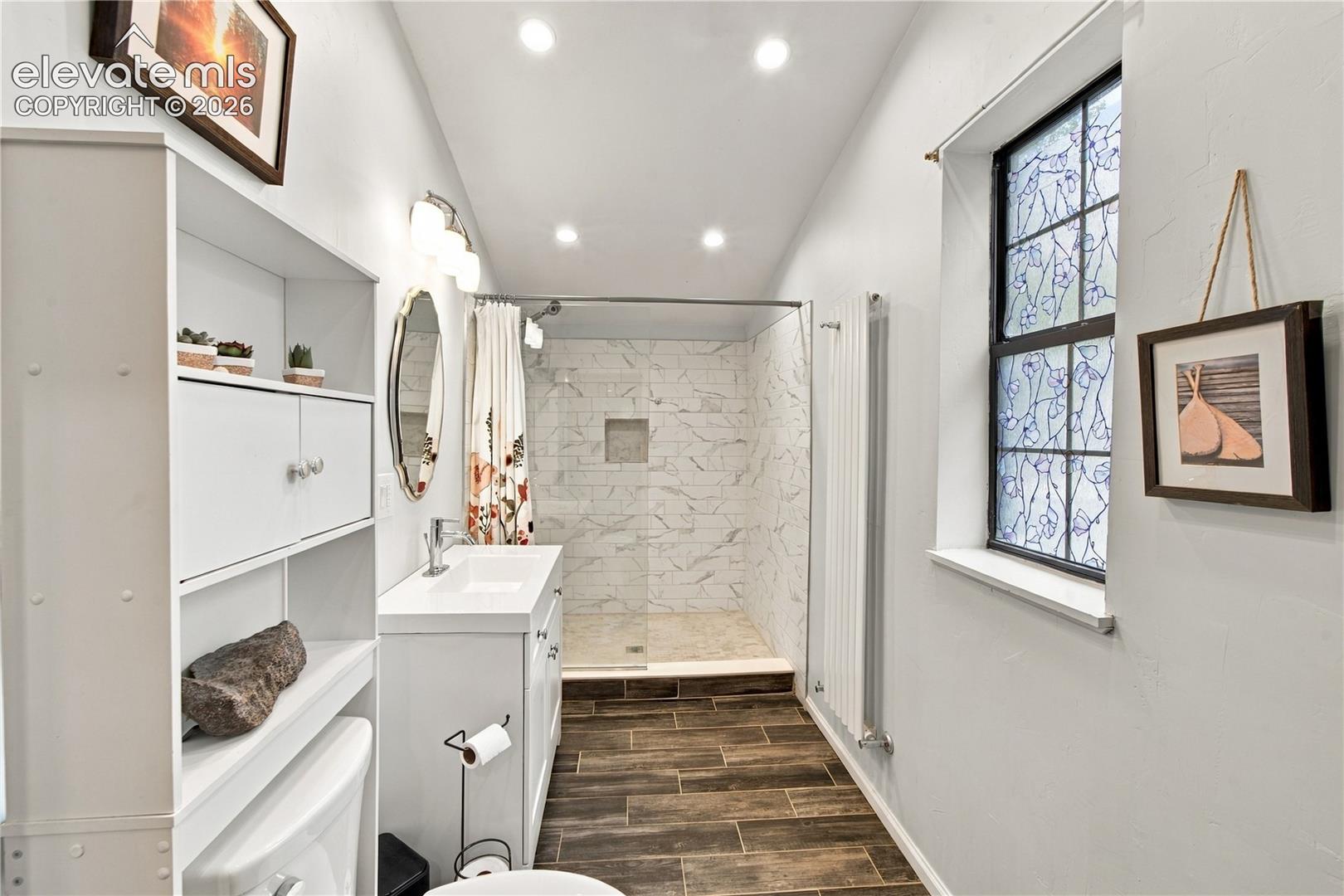 Walk-in shower with stone-pattern tile surround and built-in recessed niche, wood-finish tile flooring, white vanity with rectangular sink, recessed ceiling lighting, and decorative privacy window
