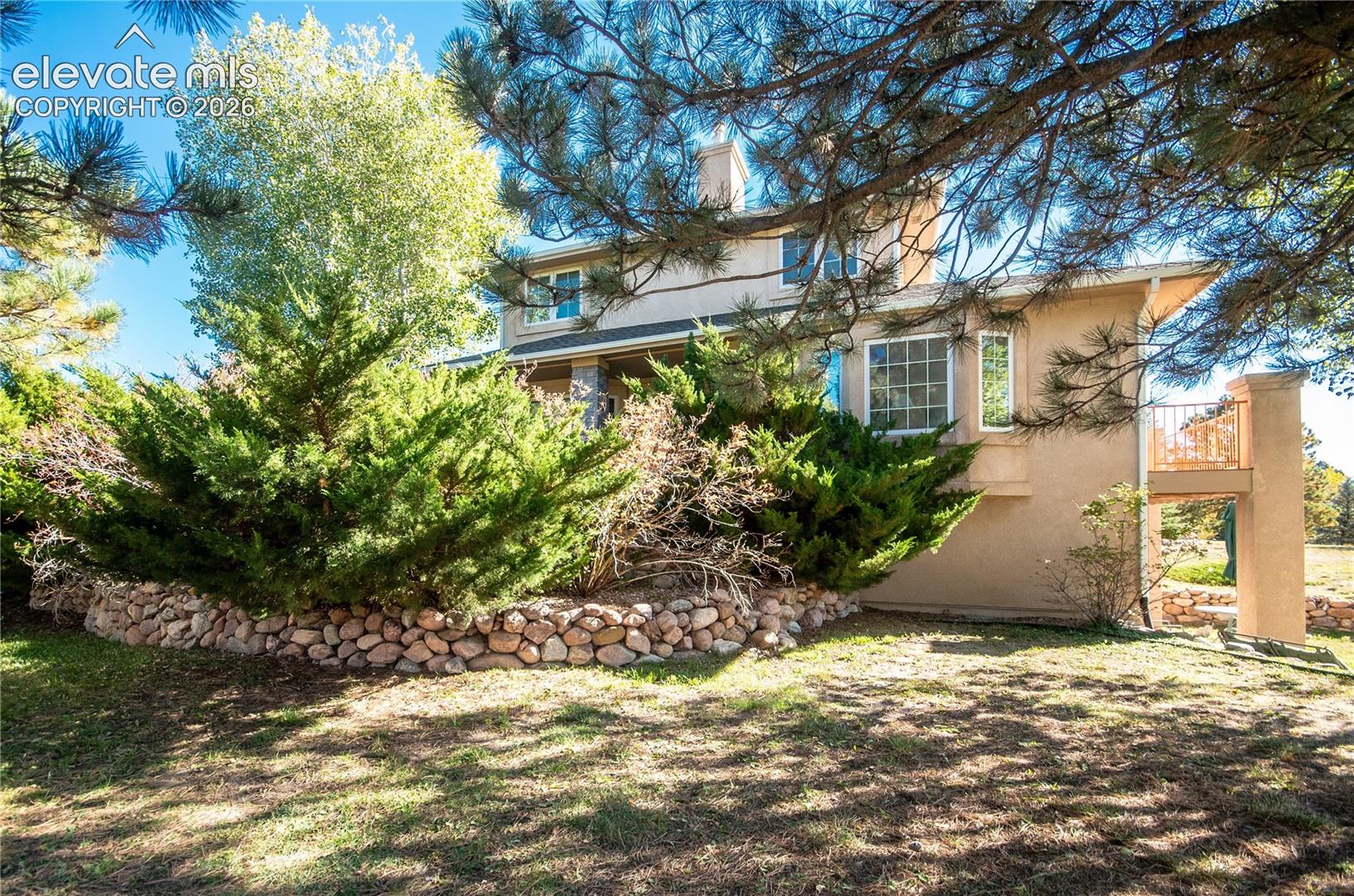 Stucco exterior with multiple windows, stone retaining wall, and mature landscaping