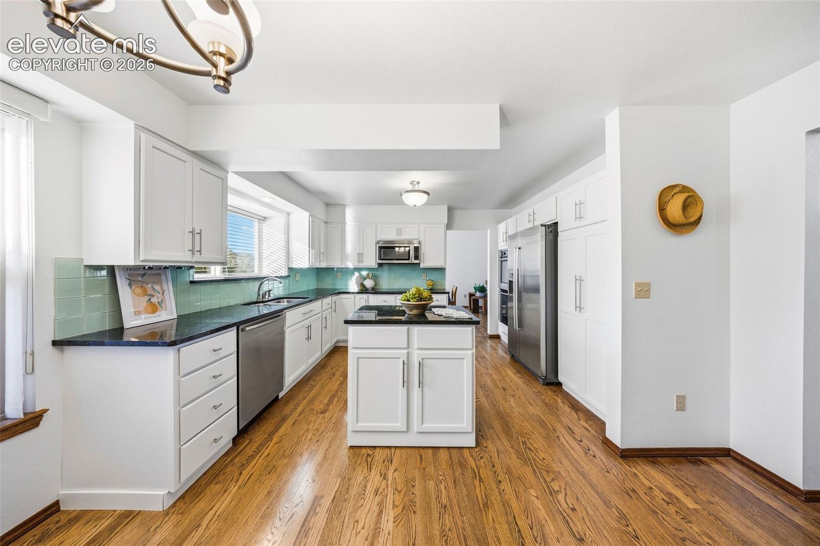 Kitchen featuring white shaker-style cabinetry, dark stone countertops, a central island with an integrated cooktop, wood-finish flooring, and stainless steel appliances