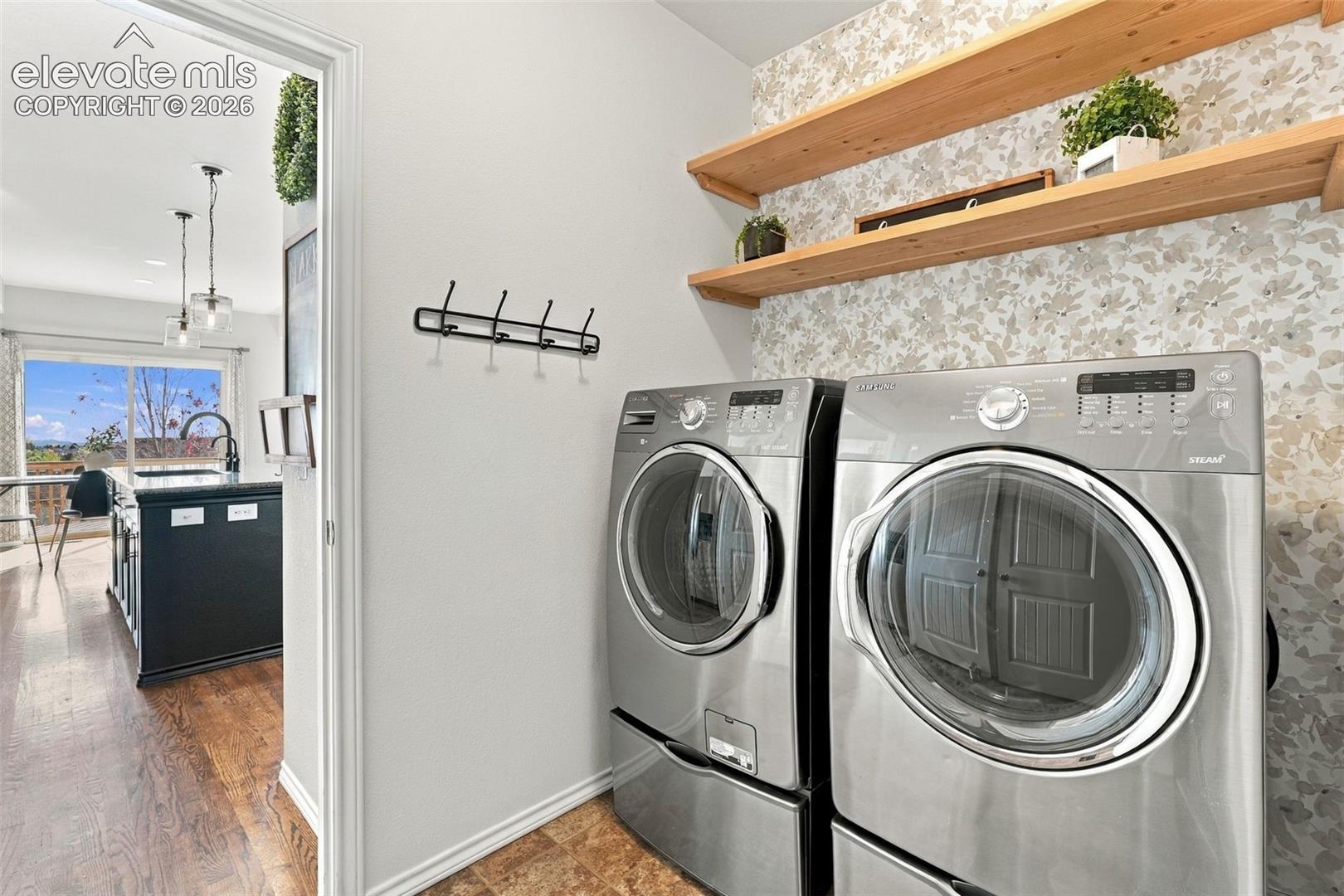 Laundry space featuring stainless steel appliances, patterned wall treatment, dual floating natural-finish wood shelves, and a wall-mounted hook rack