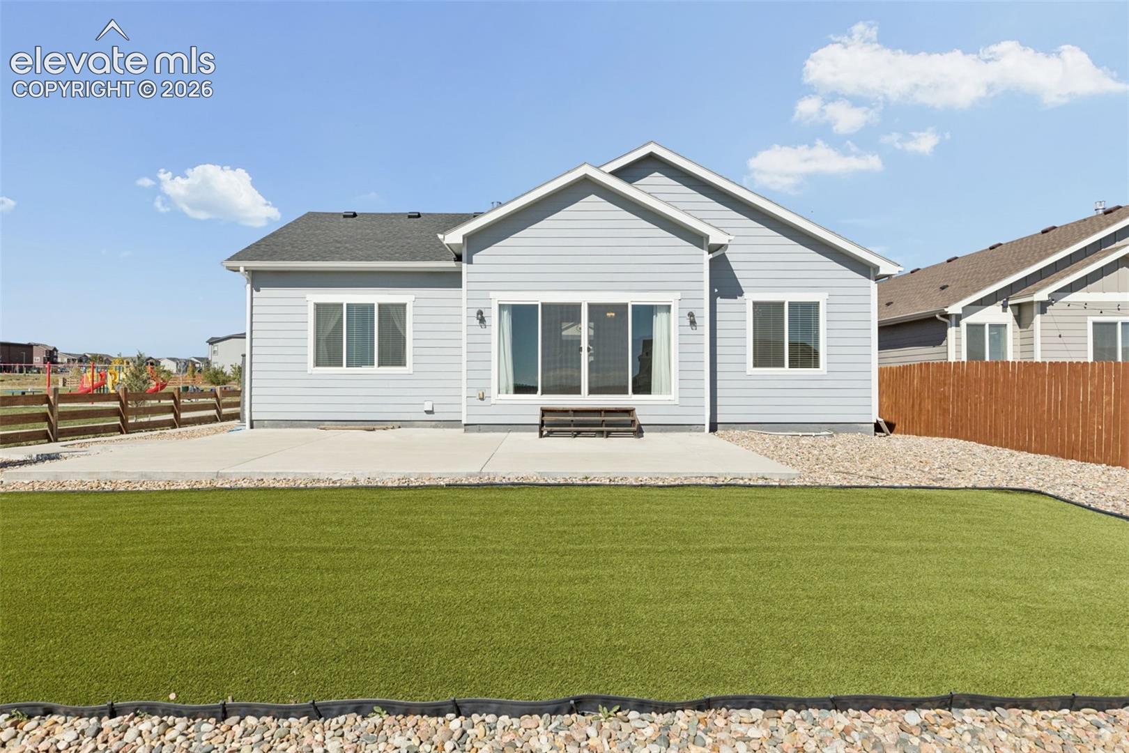 Rear exterior featuring light blue horizontal lap siding, a concrete patio, sliding glass doors, synthetic turf lawn, and perimeter wood fencing