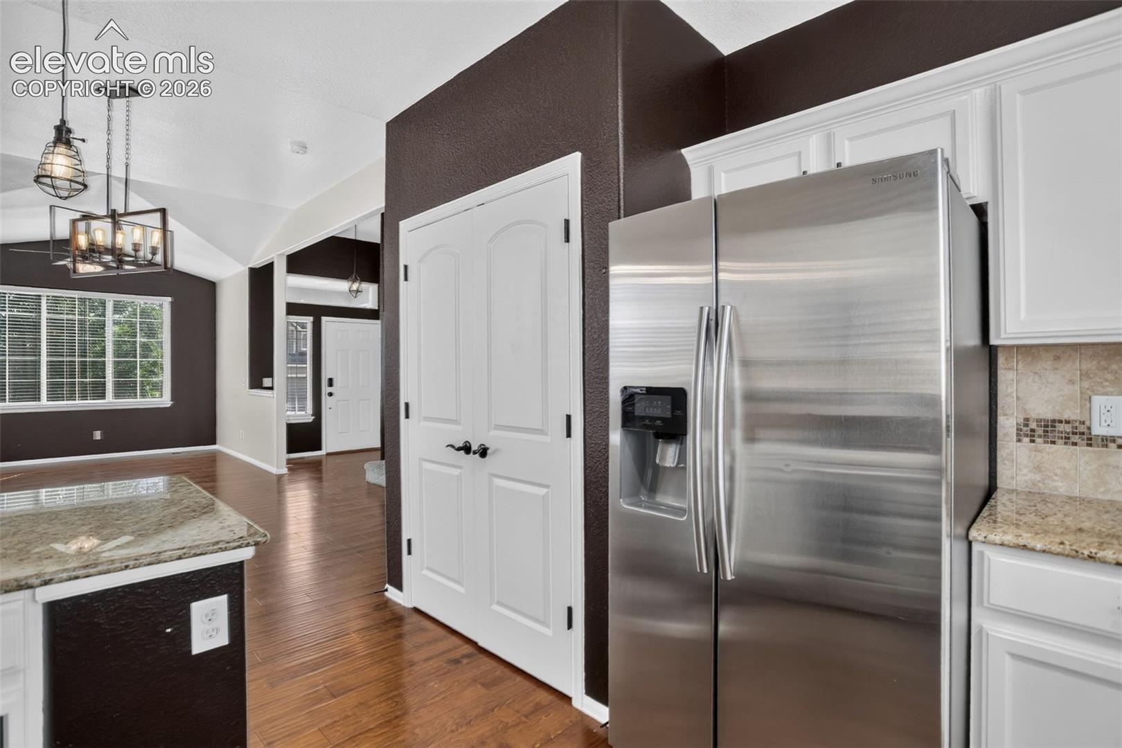 Stainless steel refrigerator, white cabinetry, light granite-style countertops, mosaic tile backsplash, and wood-finish flooring