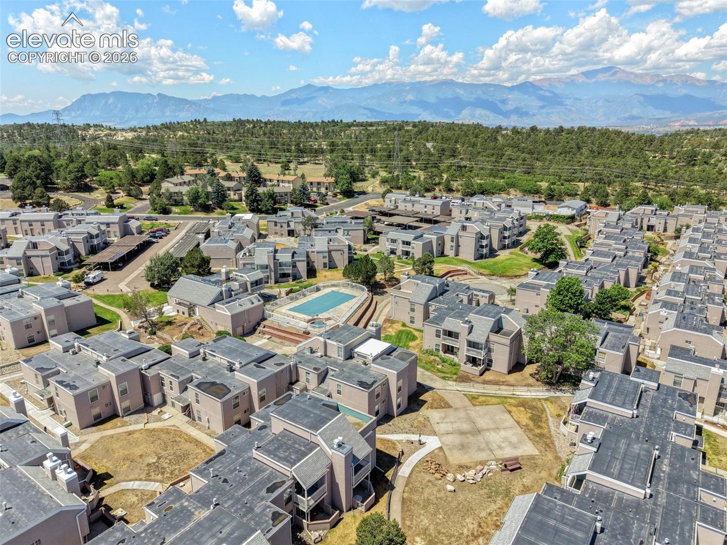 Aerial perspective featuring multi-unit residential buildings, an outdoor swimming pool, paved pedestrian walkways, and expansive mountain scenery