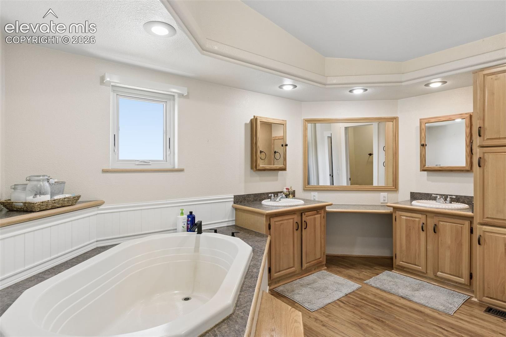 Oval soaking tub with a solid-surface surround, dual-sink wooden vanity with stone countertops, recessed lighting, and wood-finish flooring