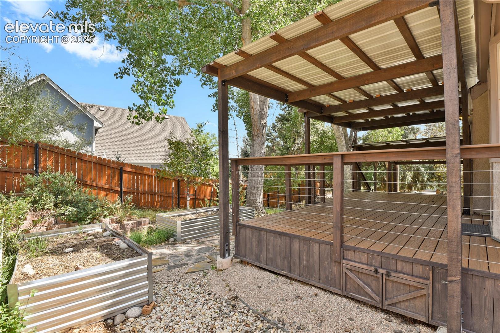 Elevated wooden deck featuring a covered pergola, cable railing system, and integrated under-deck storage cabinets, complemented by corrugated metal garden beds and natural stone hardscaping