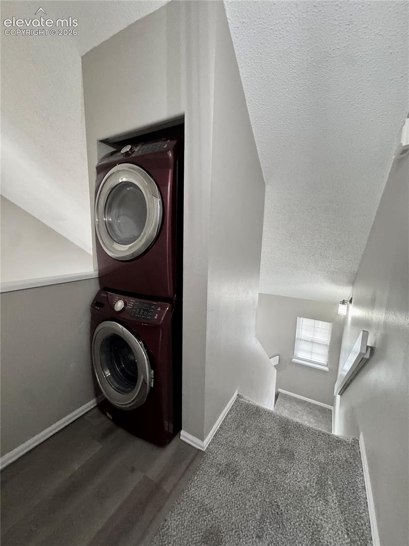 Stackable laundry appliances within an alcove, featuring wood-finish flooring and light gray carpeting