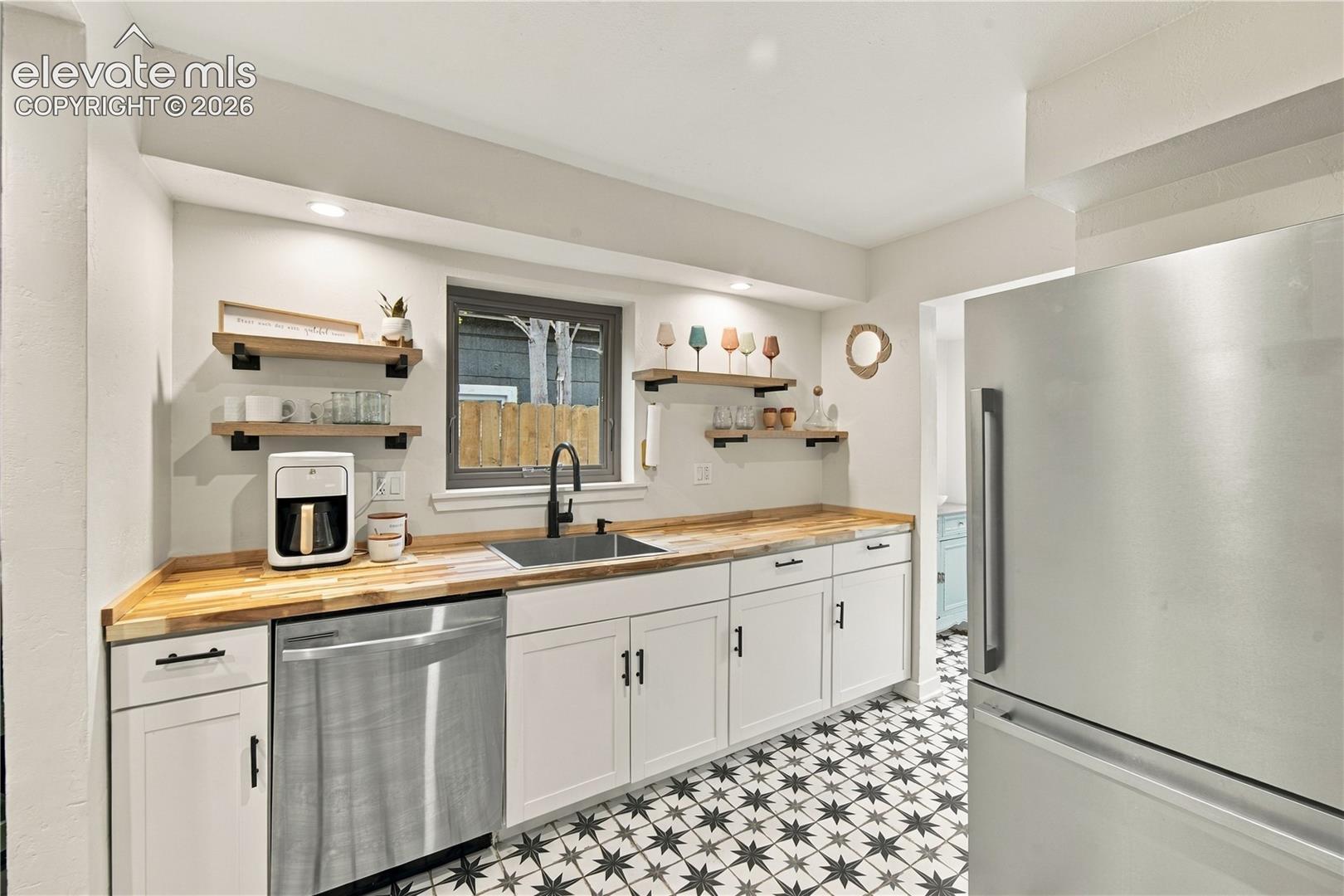 Kitchen featuring white cabinetry with matte black hardware, wood-finish countertops, geometric patterned tile flooring, stainless steel appliances, and wall-mounted wooden shelving