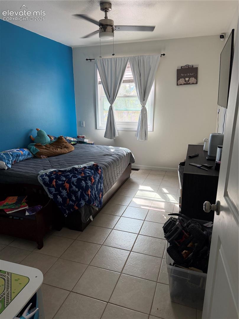 Recessed window with drapes, ceiling fan with light fixture, light-toned tile flooring, and a wall-mounted television