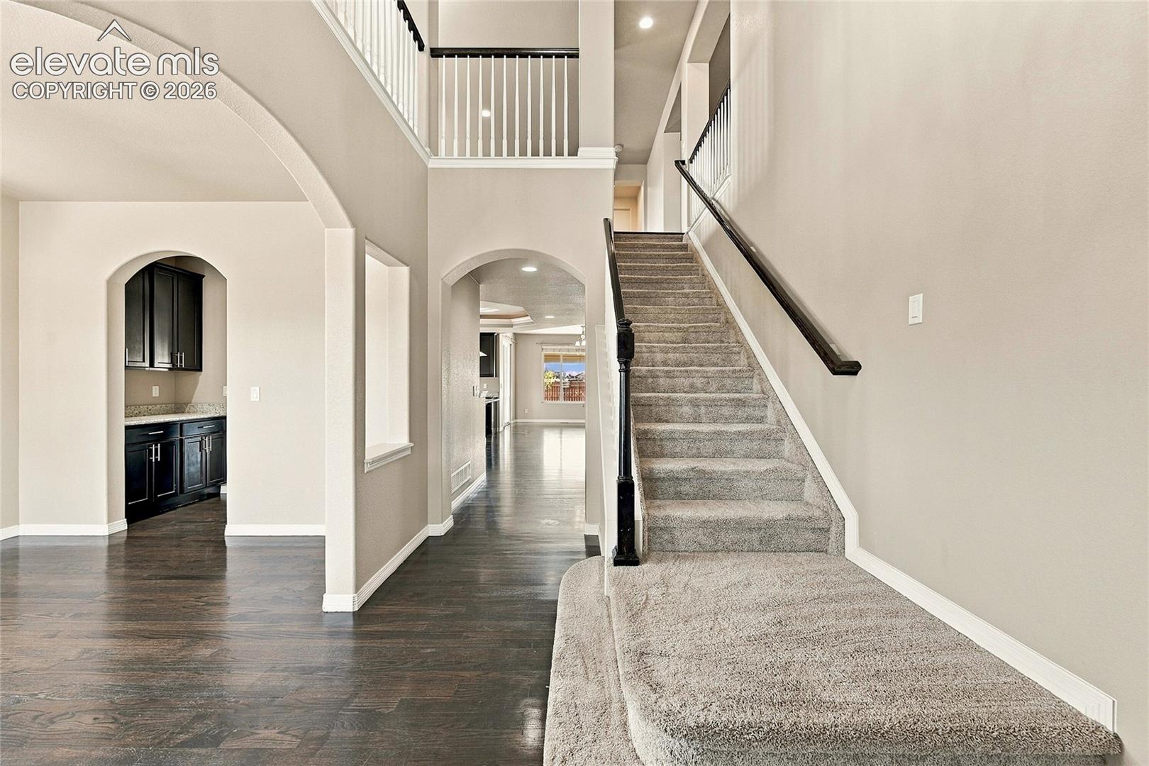 Two-story foyer featuring dark wood-finish flooring, a carpeted staircase with a dark wood handrail, arched architectural detailing, and recessed lighting