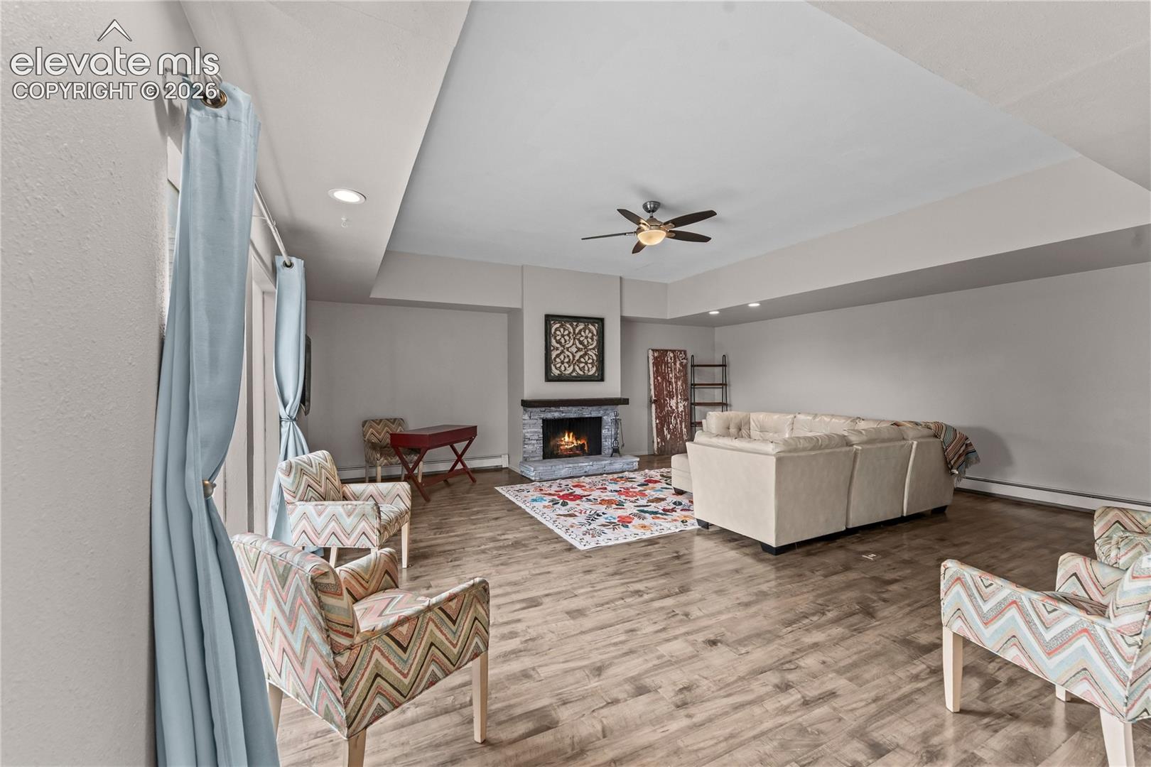 Living area featuring wood-finish flooring, a tray ceiling with recessed lighting, a ceiling fan, and a stone-surround fireplace with a wooden mantel