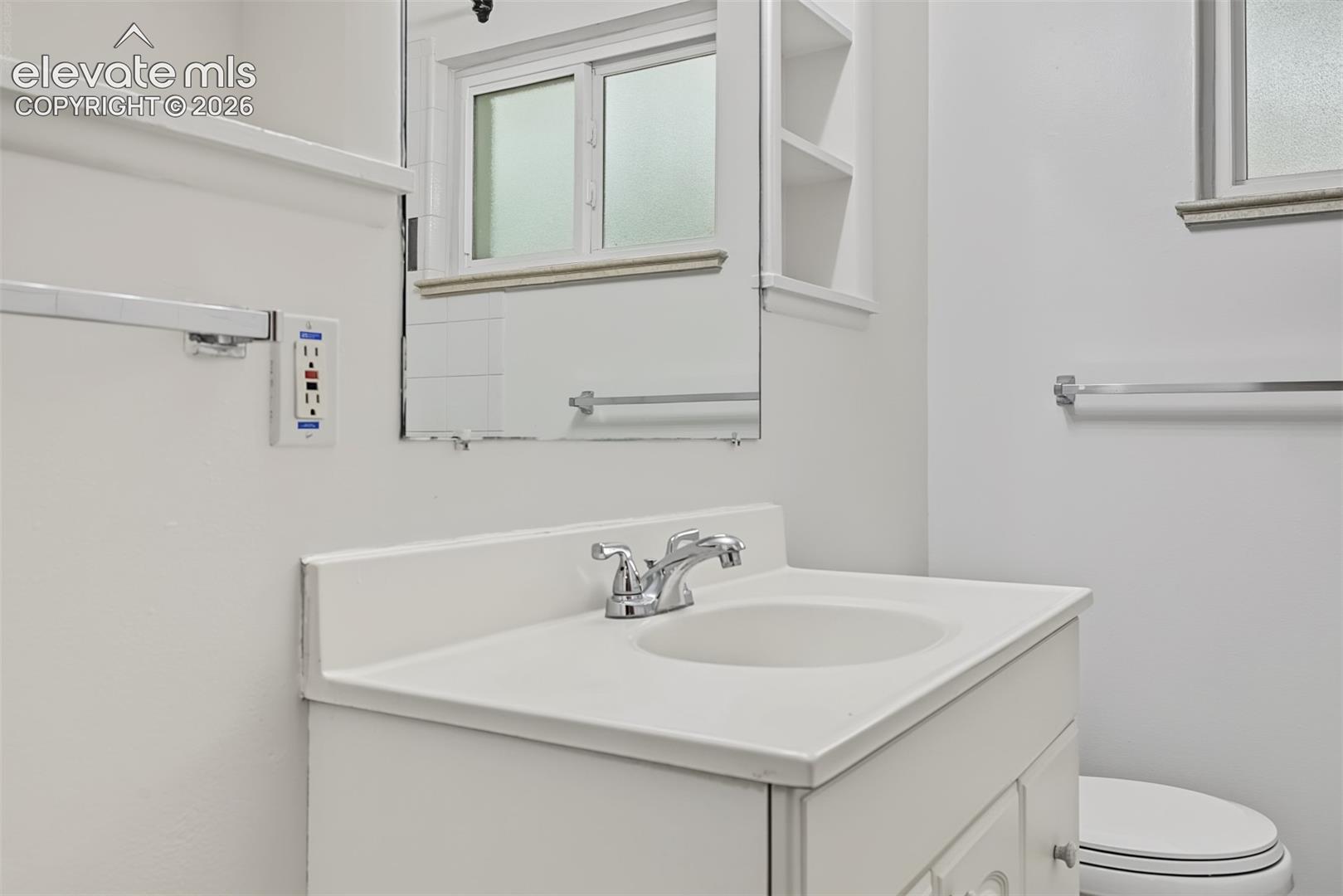 Vanity featuring a white countertop with integrated sink and chrome faucet, rectangular wall mirror with built-in shelving, and polished chrome towel bars