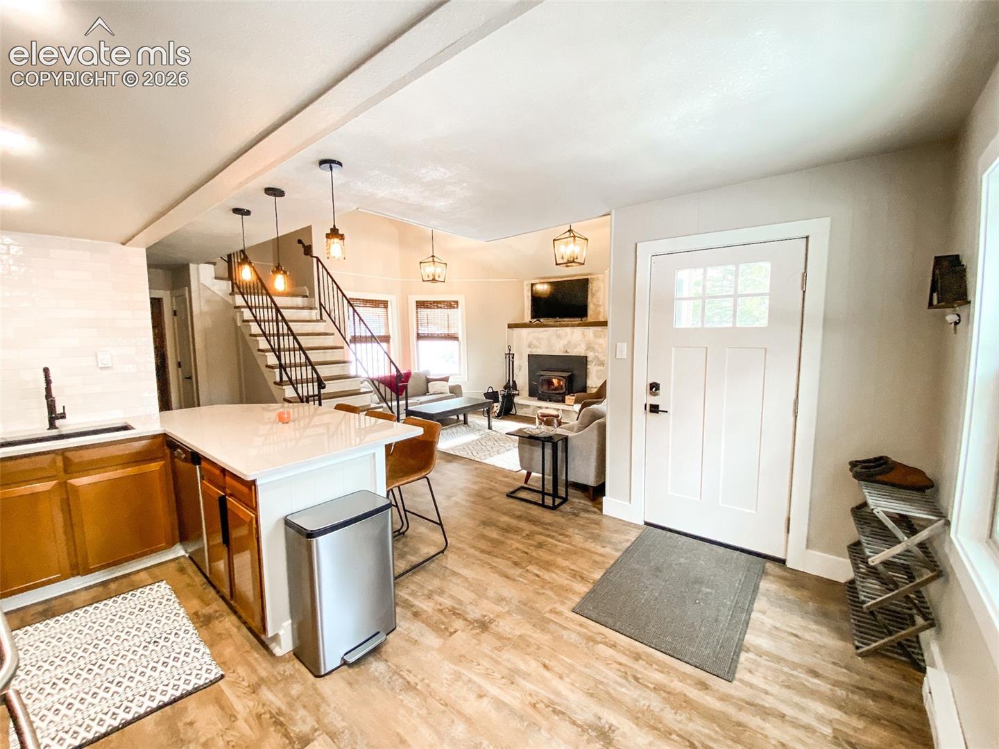 Open-concept living space featuring wood-finish flooring, a kitchen island with seating, a staircase with a black metal railing, a stone fireplace, and a white paneled entry door with glass inserts